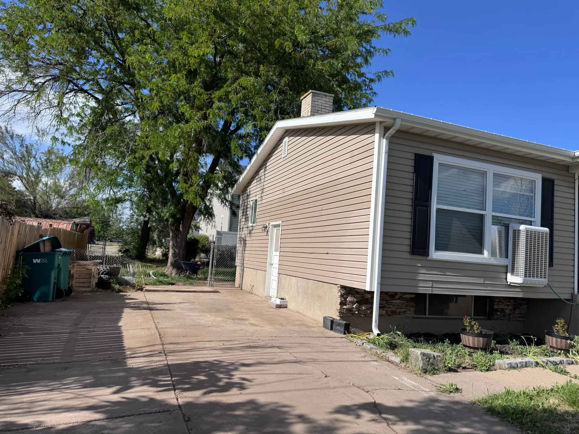 Tan house with a driveway. Tree in the background. Green trash bins to the left. Sunny day.