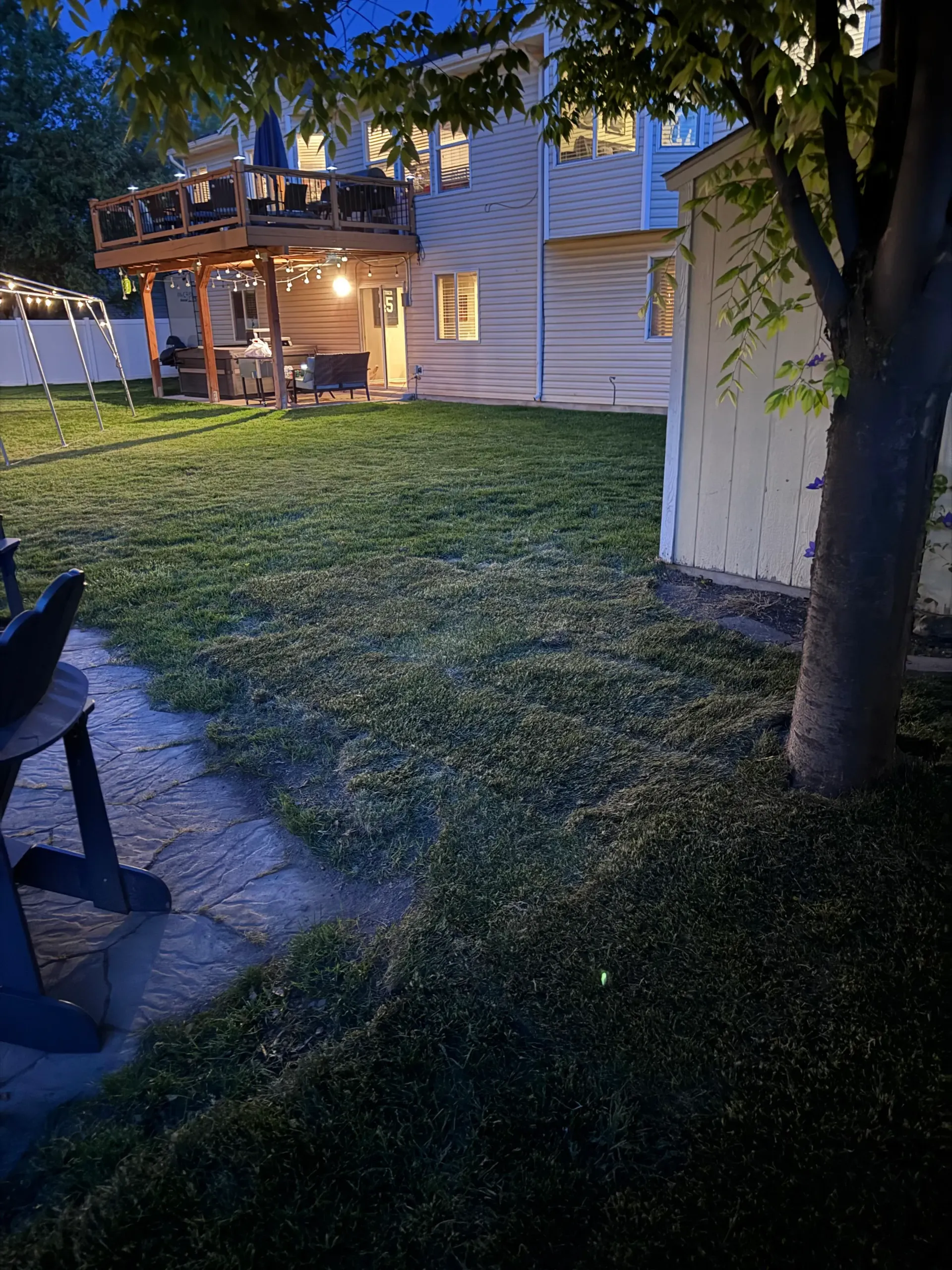 Backyard at dusk with a multi-level deck, lush green lawn, tree, and shed; house in the background.