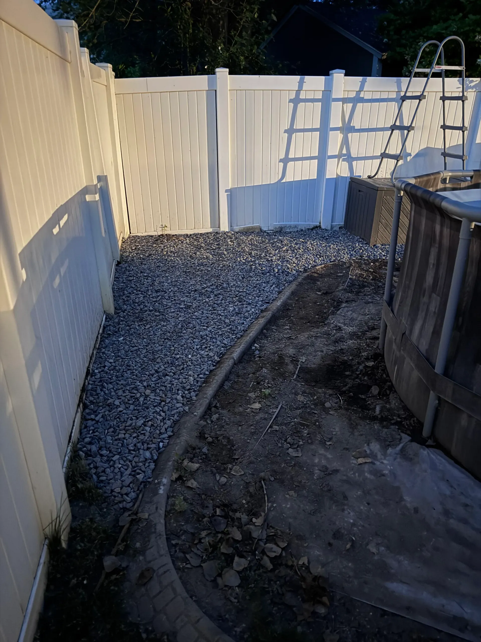 Gravel path between white fences, leading to a gate and an above-ground pool. Ladder leans on the far fence.