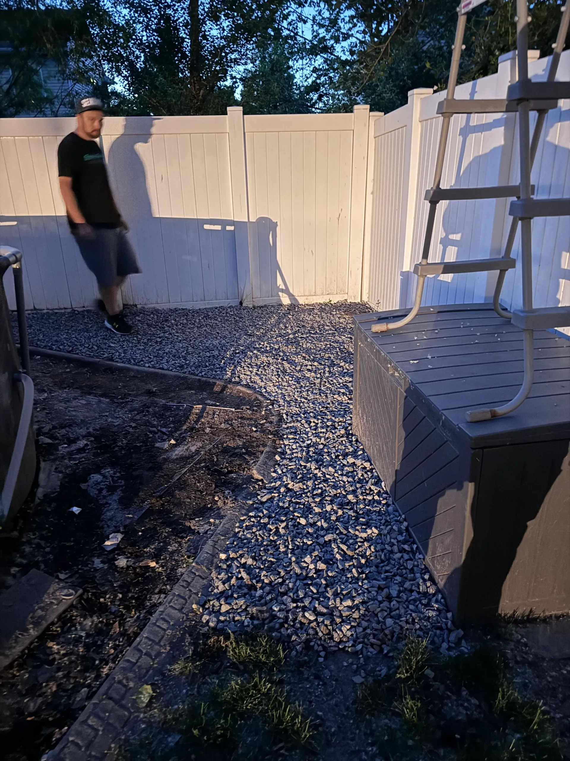 Man in shorts and t-shirt stands near gravel path and white fence next to a pool ladder.