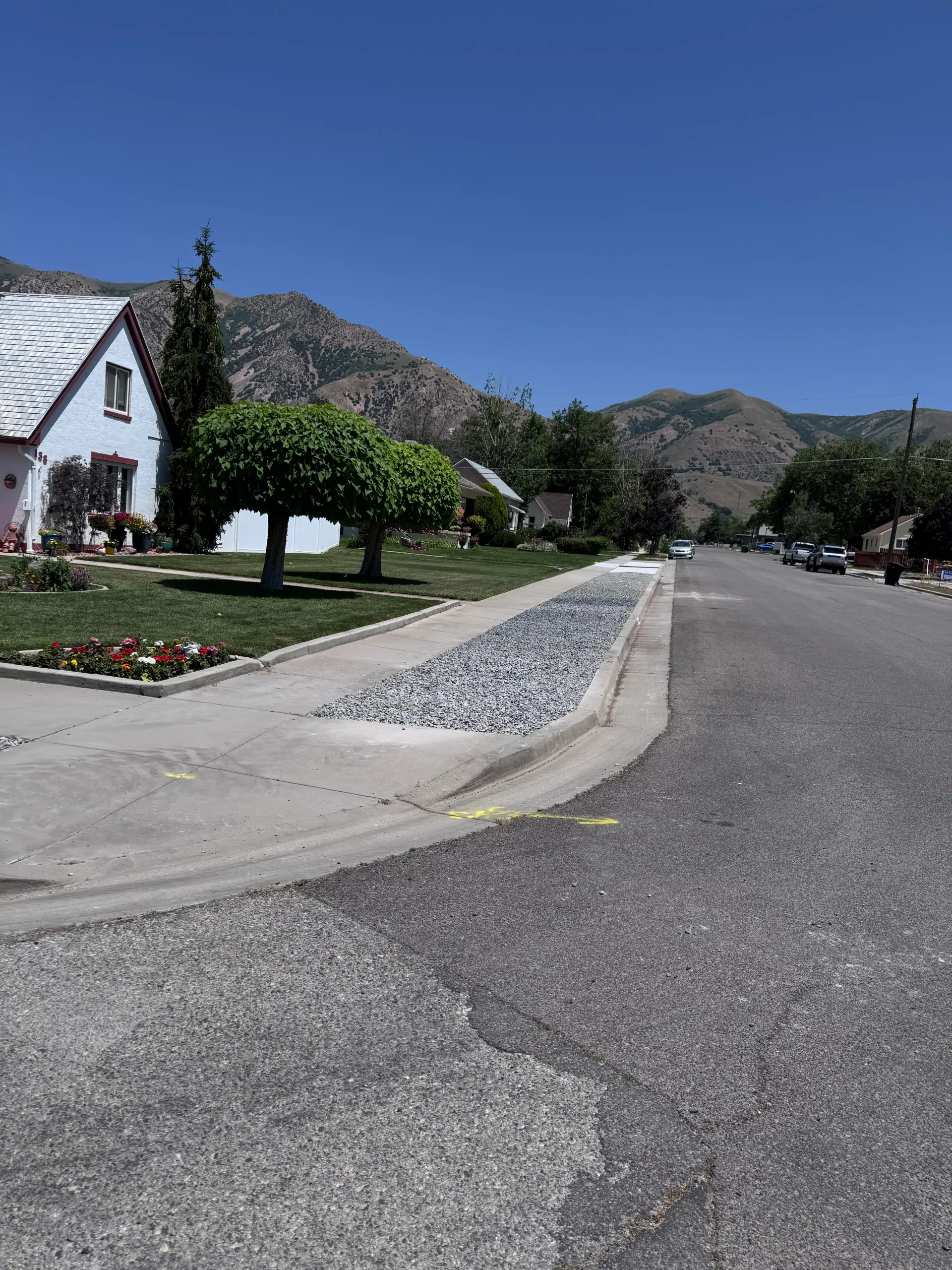 Street with stone-filled gutter, houses, trees, and mountains under a blue sky.