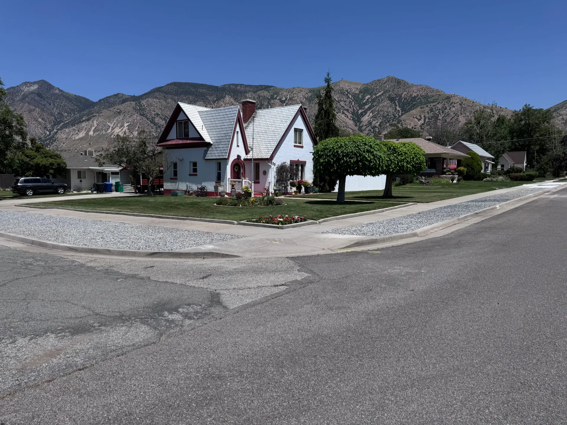Small blue house with red trim, gravel drive, street corner, mountains in background, clear sky.