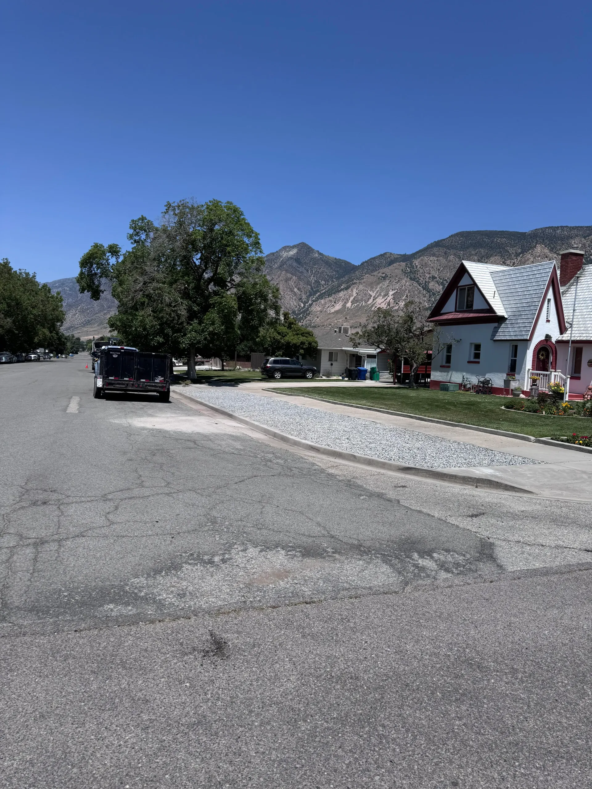 Paved road with parked vehicles, houses, and mountains under a clear blue sky.