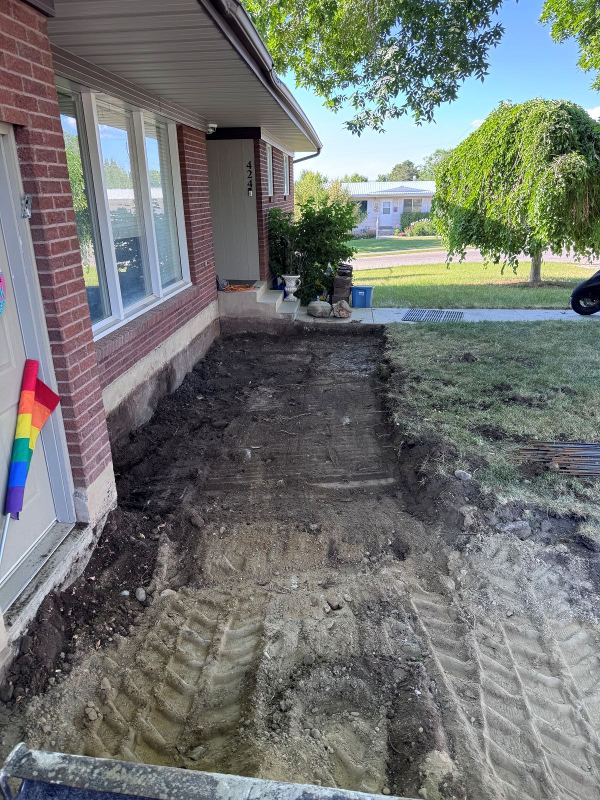 Excavated dirt bed in front of a brick house with grass and trees. A rainbow flag hangs on the door.