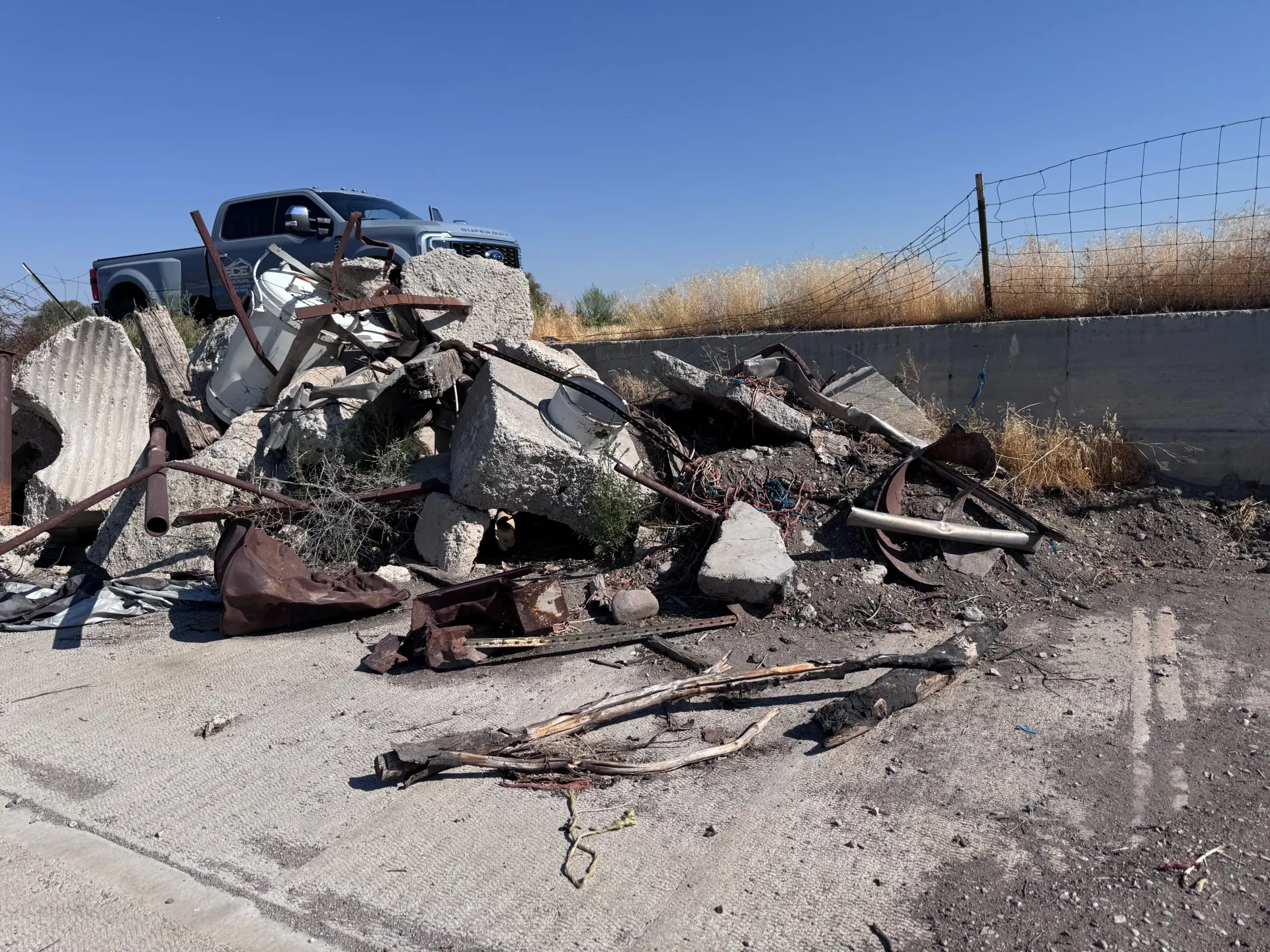 Pile of debris with a truck visible, against a barbed-wire fence and blue sky.