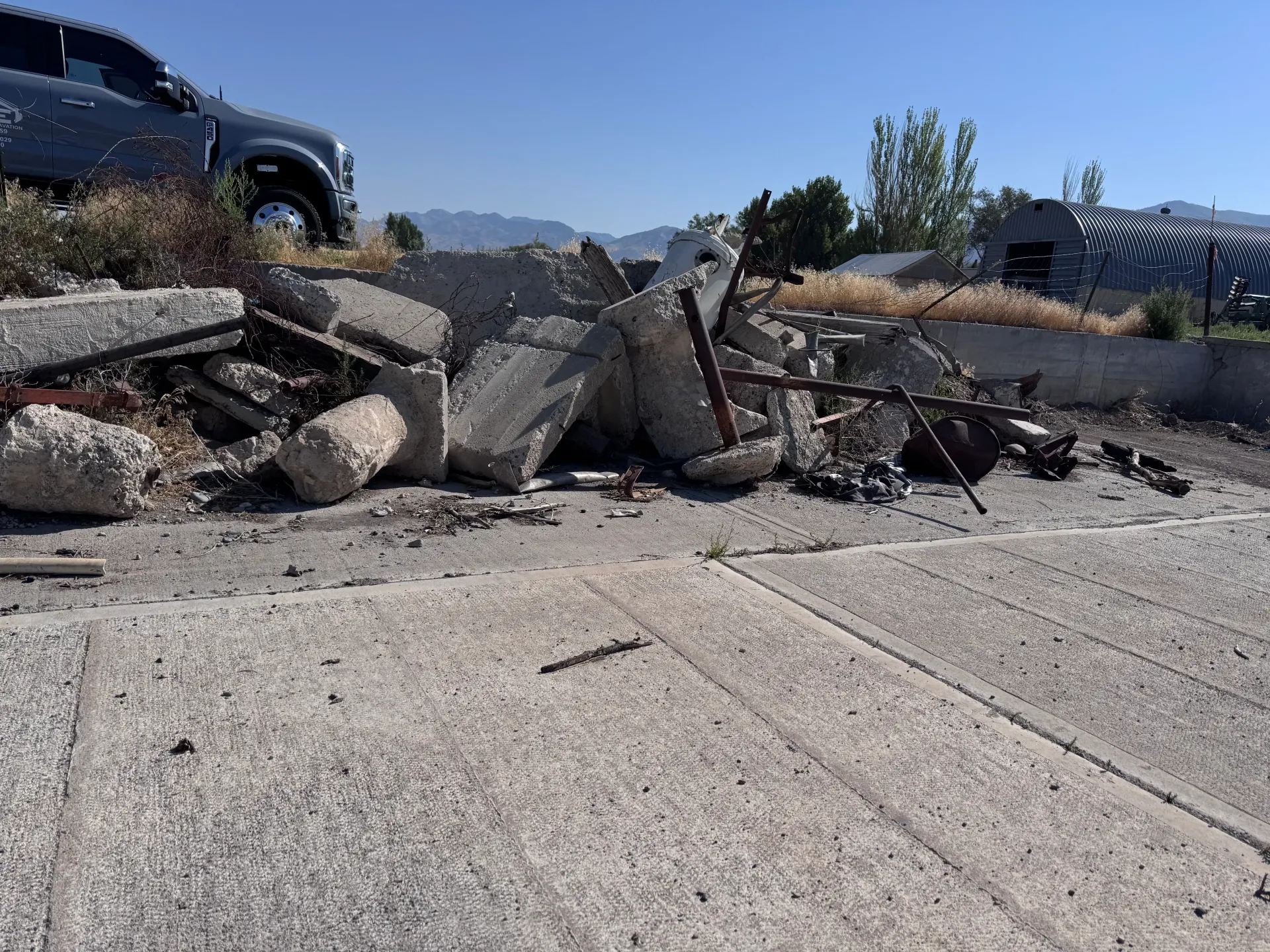 Concrete rubble and metal debris on a sidewalk next to a road, mountains in the distance.