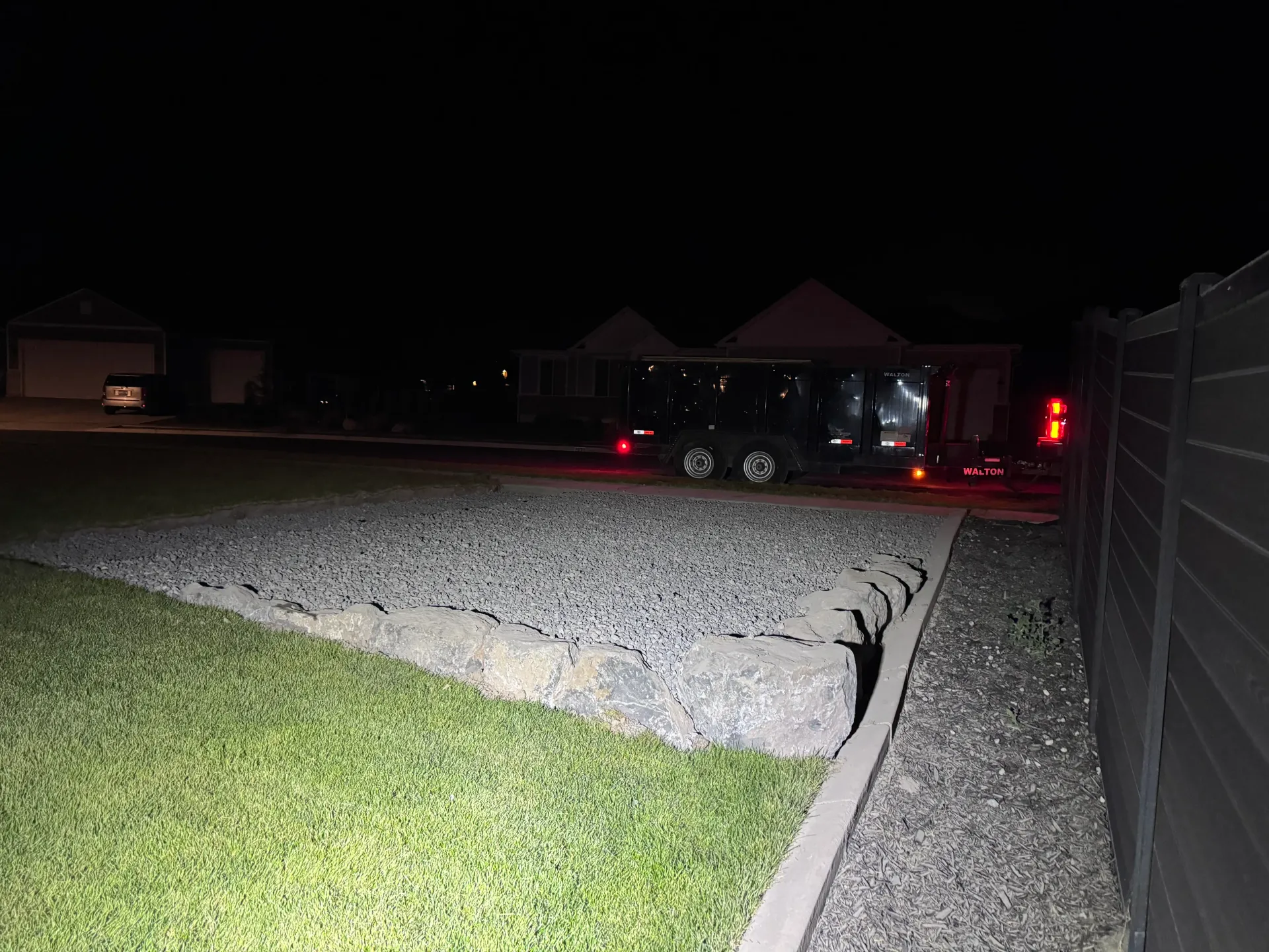 Dark night scene with a vehicle parked near a house, gravel bed in foreground, lights on.