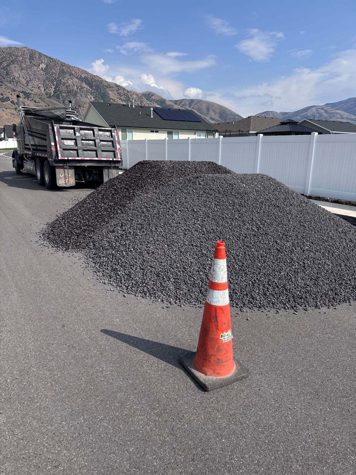 Dump truck unloading a pile of gravel on asphalt, orange traffic cone in the foreground, mountains and houses in the background.