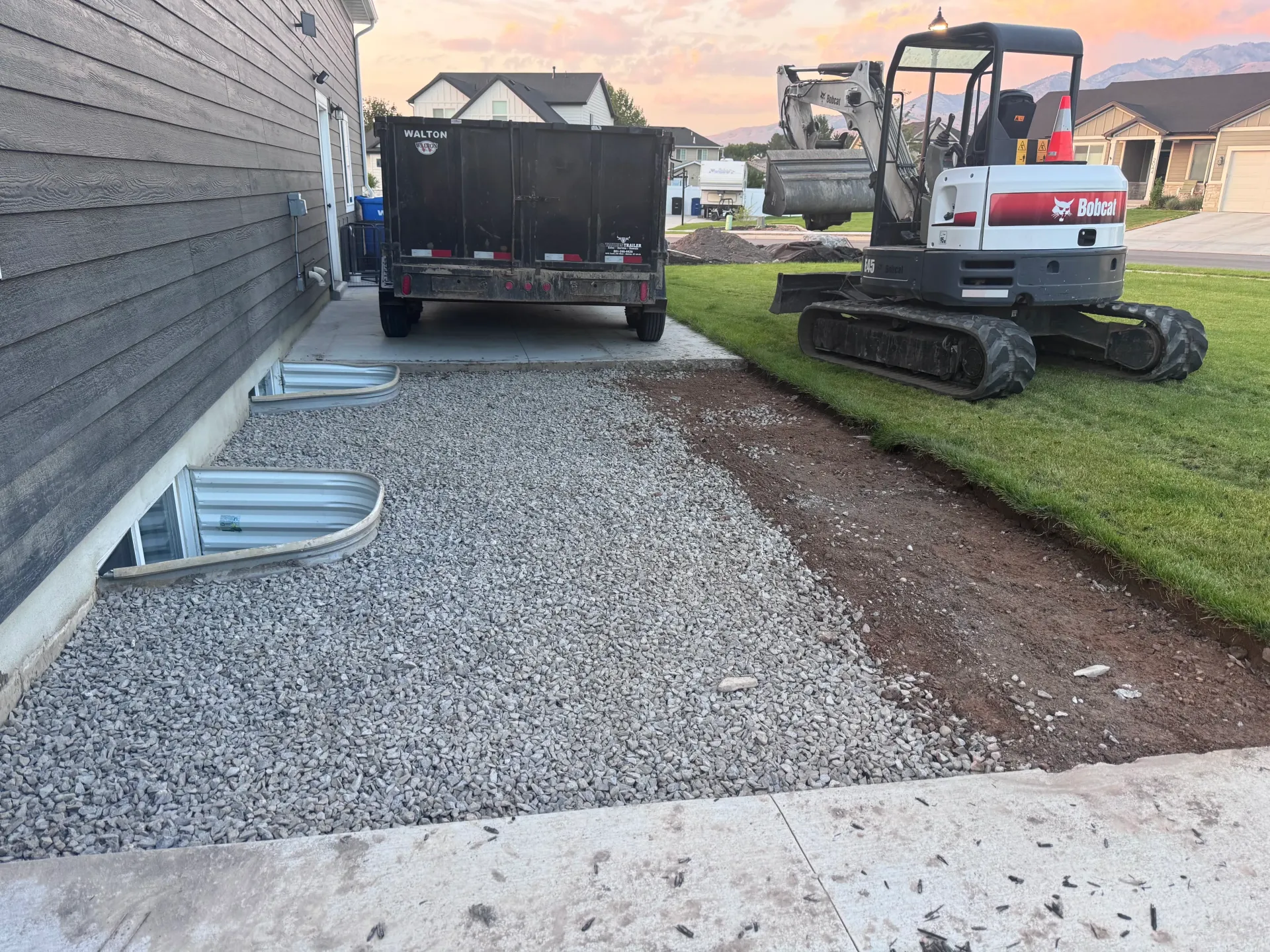 Construction site next to a house with an excavator, gravel, and a trailer. Evening light.
