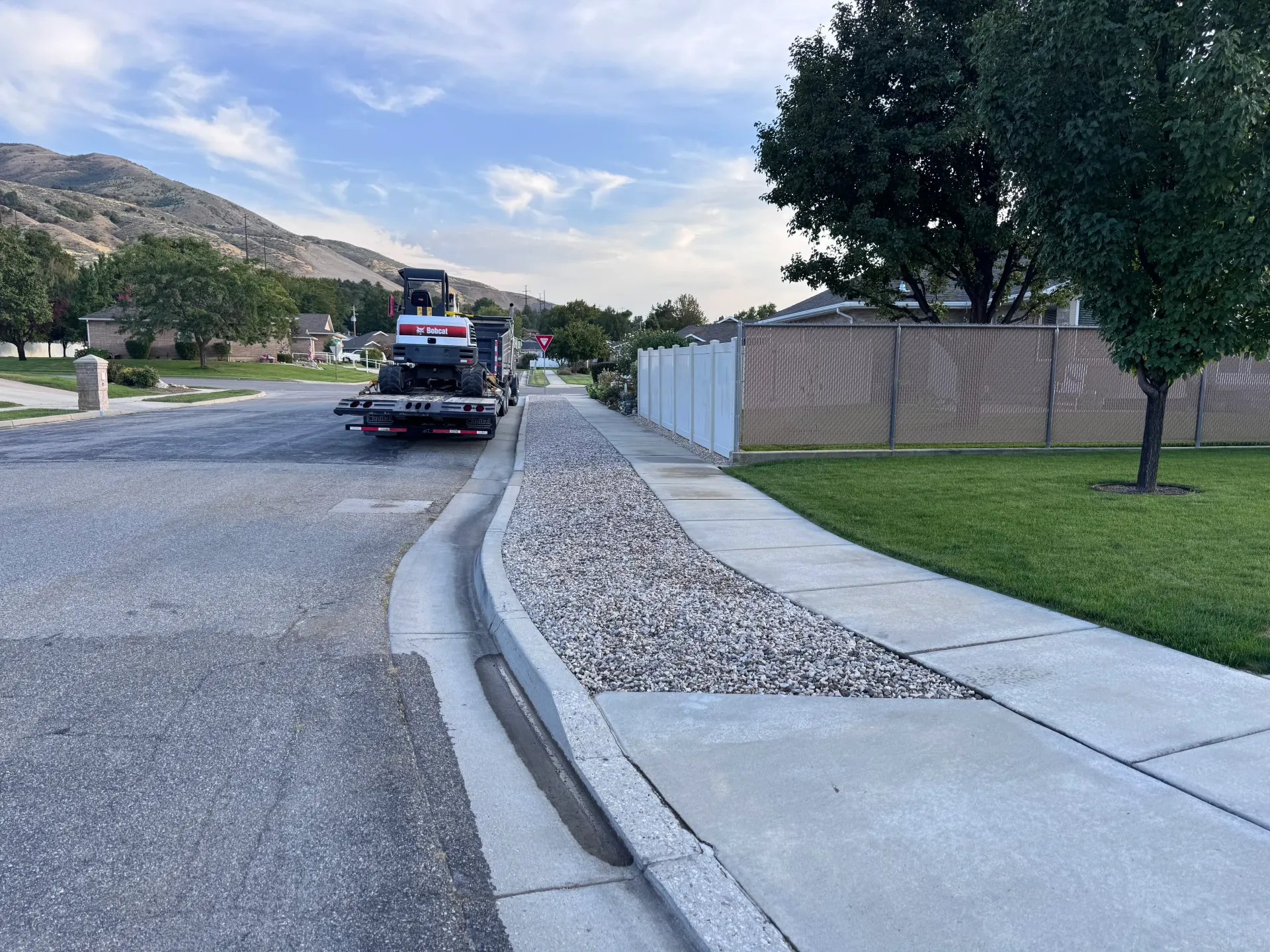 Street with gravel bed, sidewalk, and curb. A trailer is parked. Mountains in the background.