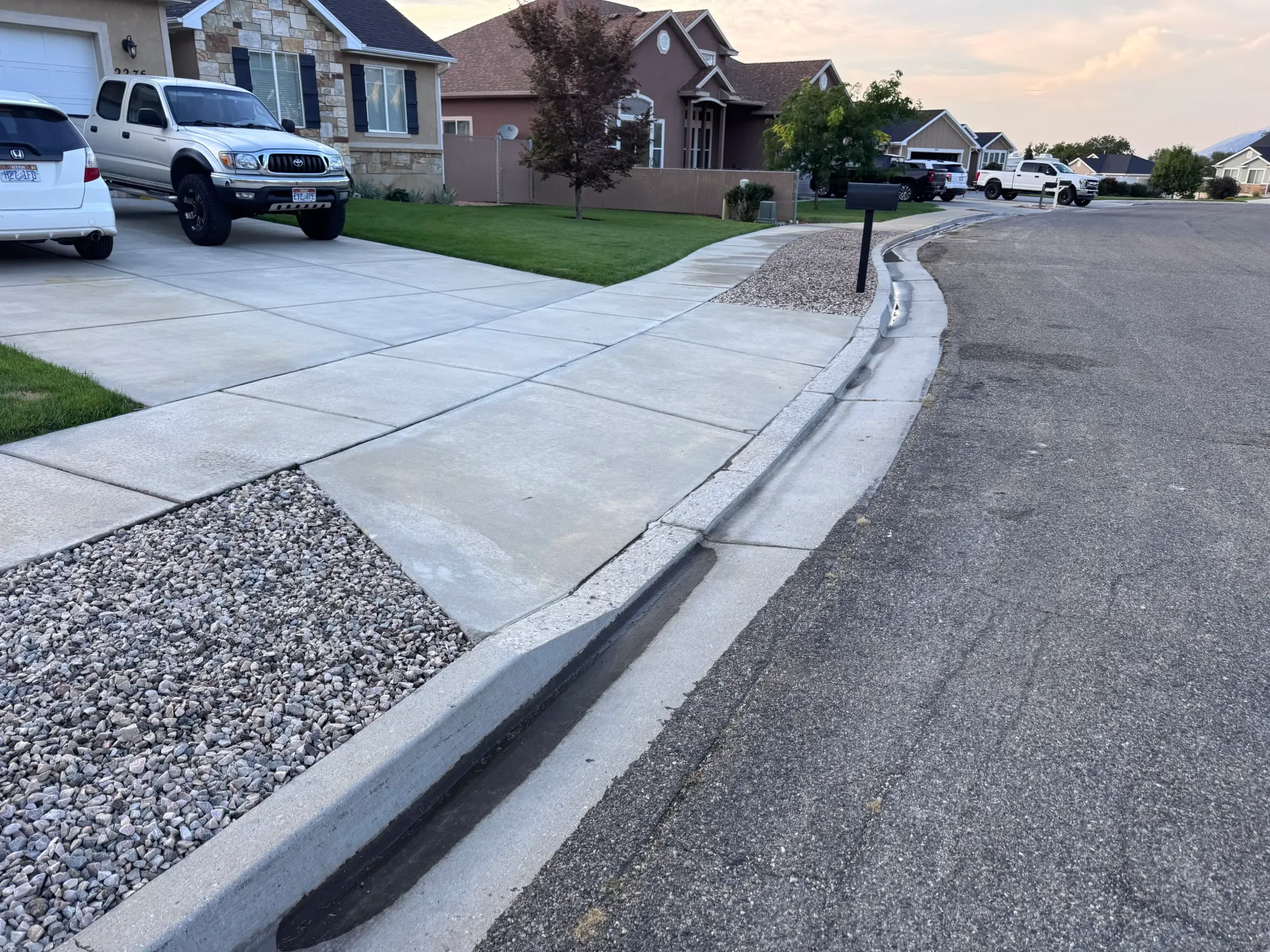 Concrete curb and gutter along a street with gravel shoulder and driveway with vehicles parked.