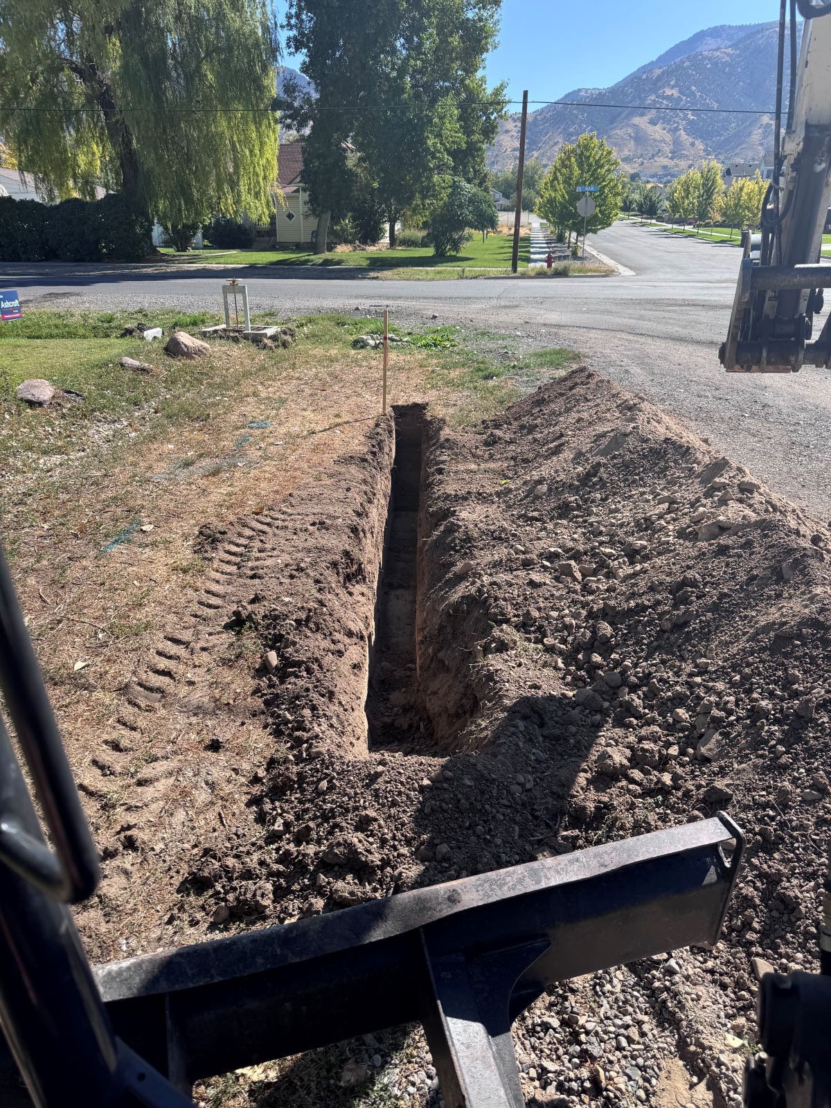 Trench dug in dirt, likely for utility lines, next to road and residential area.