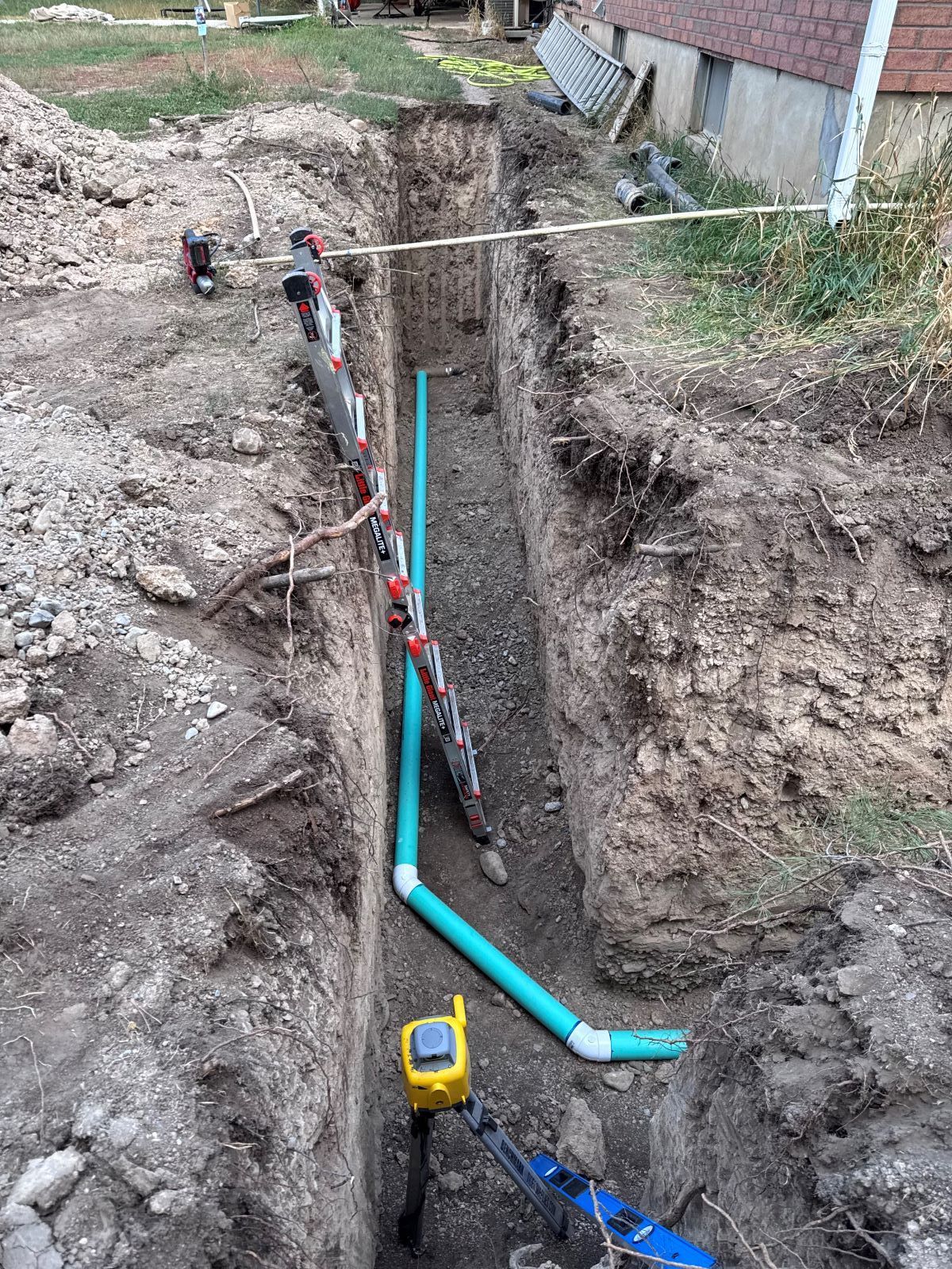 Trench dug with blue pipes installed, equipment visible, near a building.