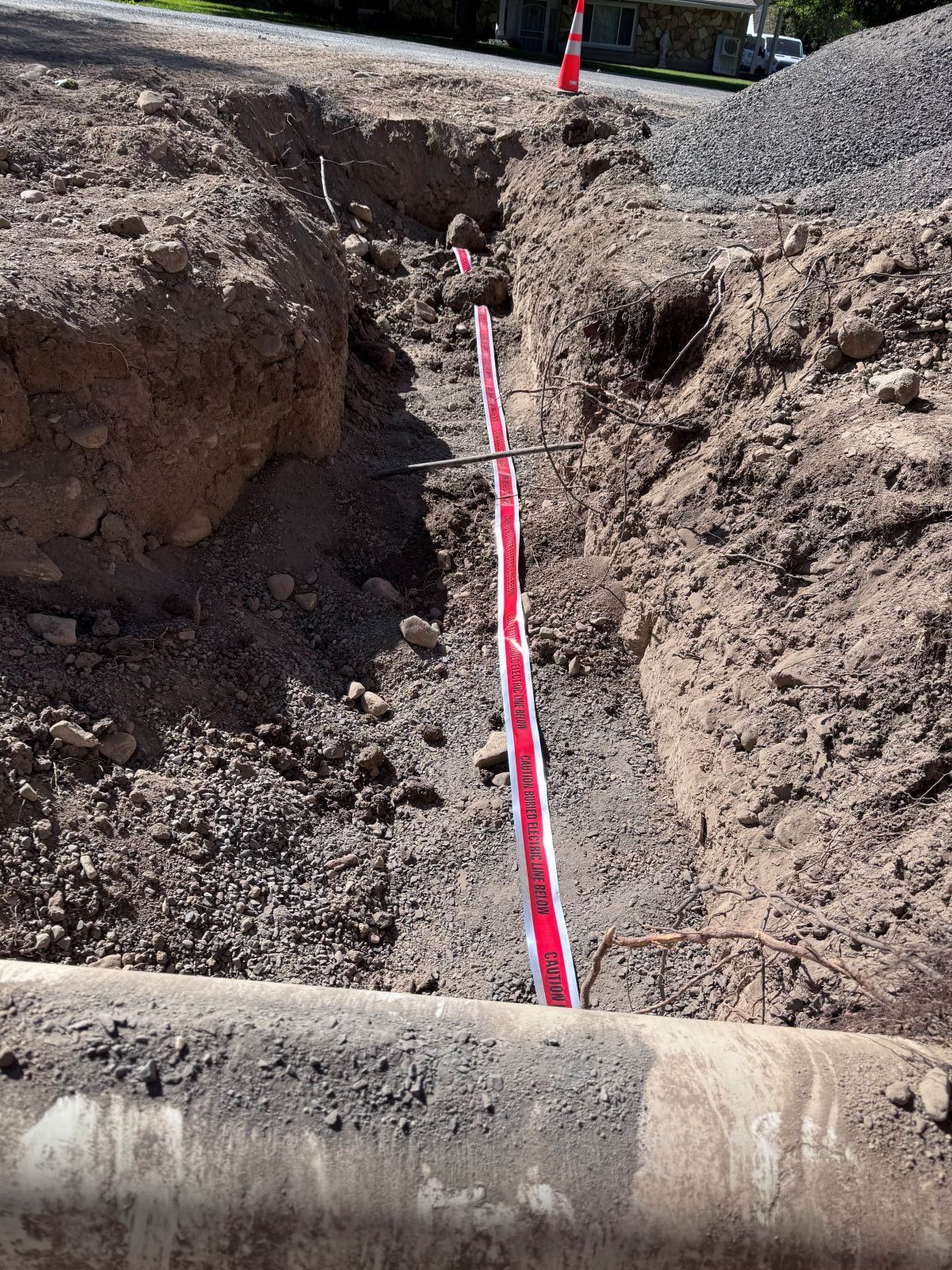 Trench in the ground with red tape, potentially for utility work. Orange cone and gravel present.