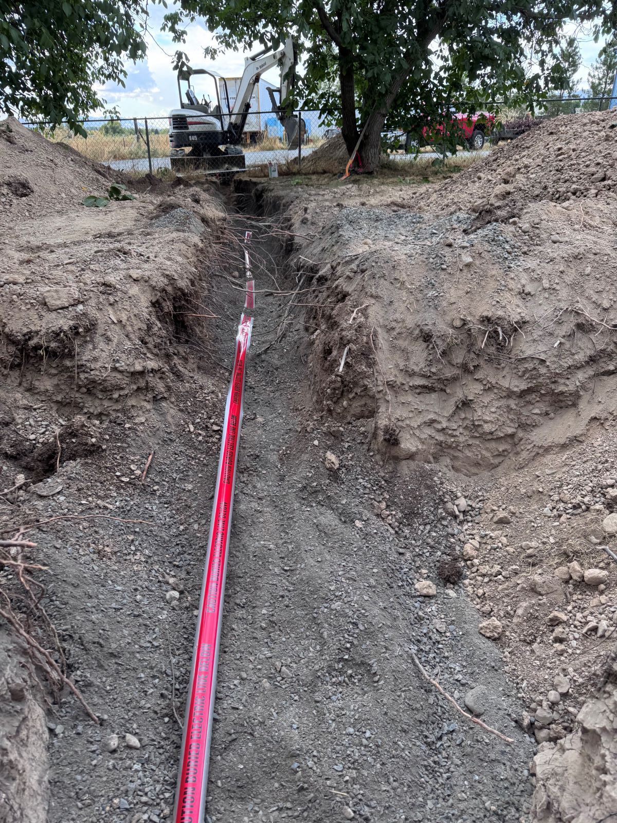 Trench with red conduit, excavated dirt, and a small backhoe in the background.