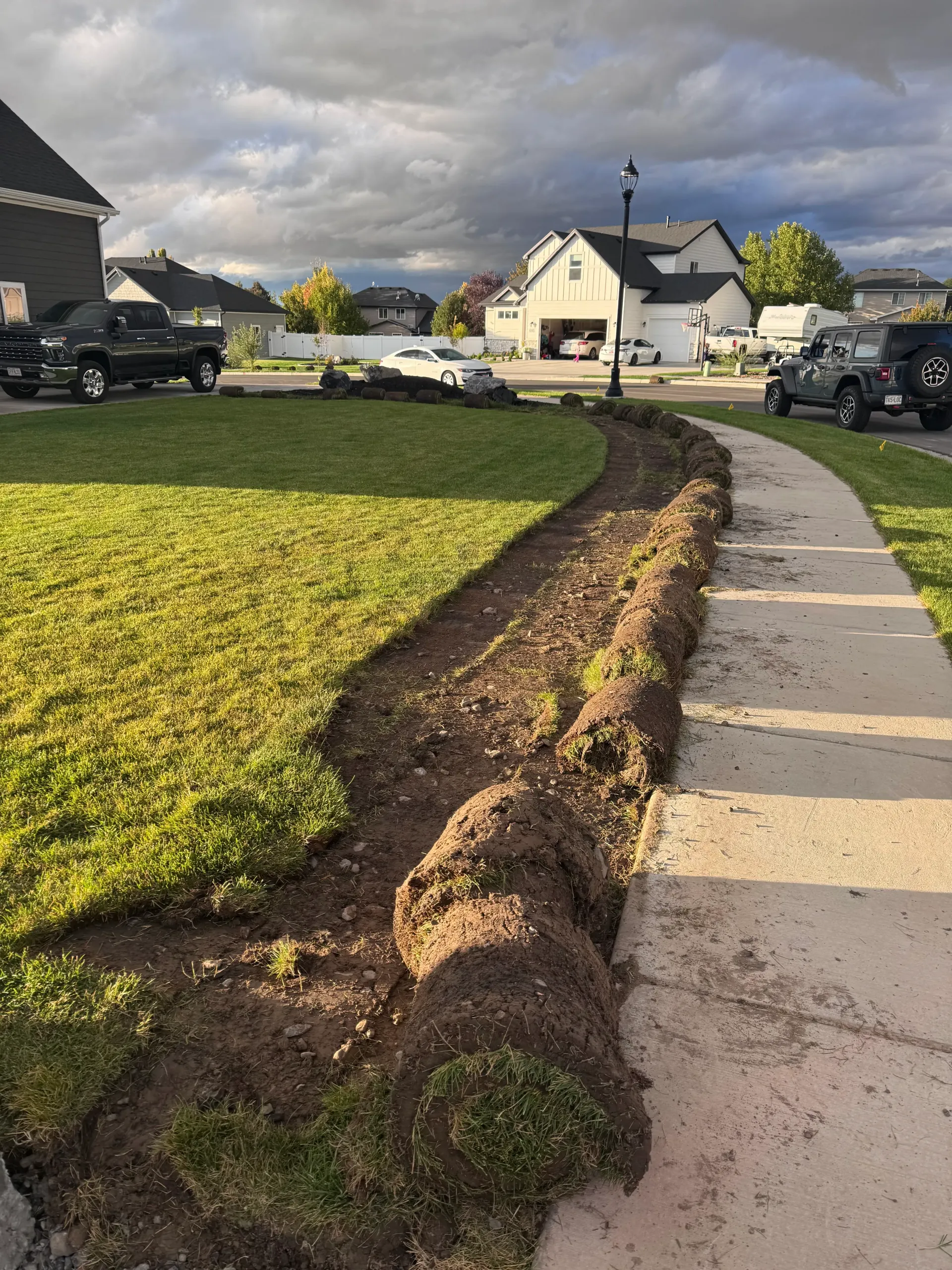 A strip of uprooted grass sits next to a sidewalk. A cloudy sky looms over a residential street.
