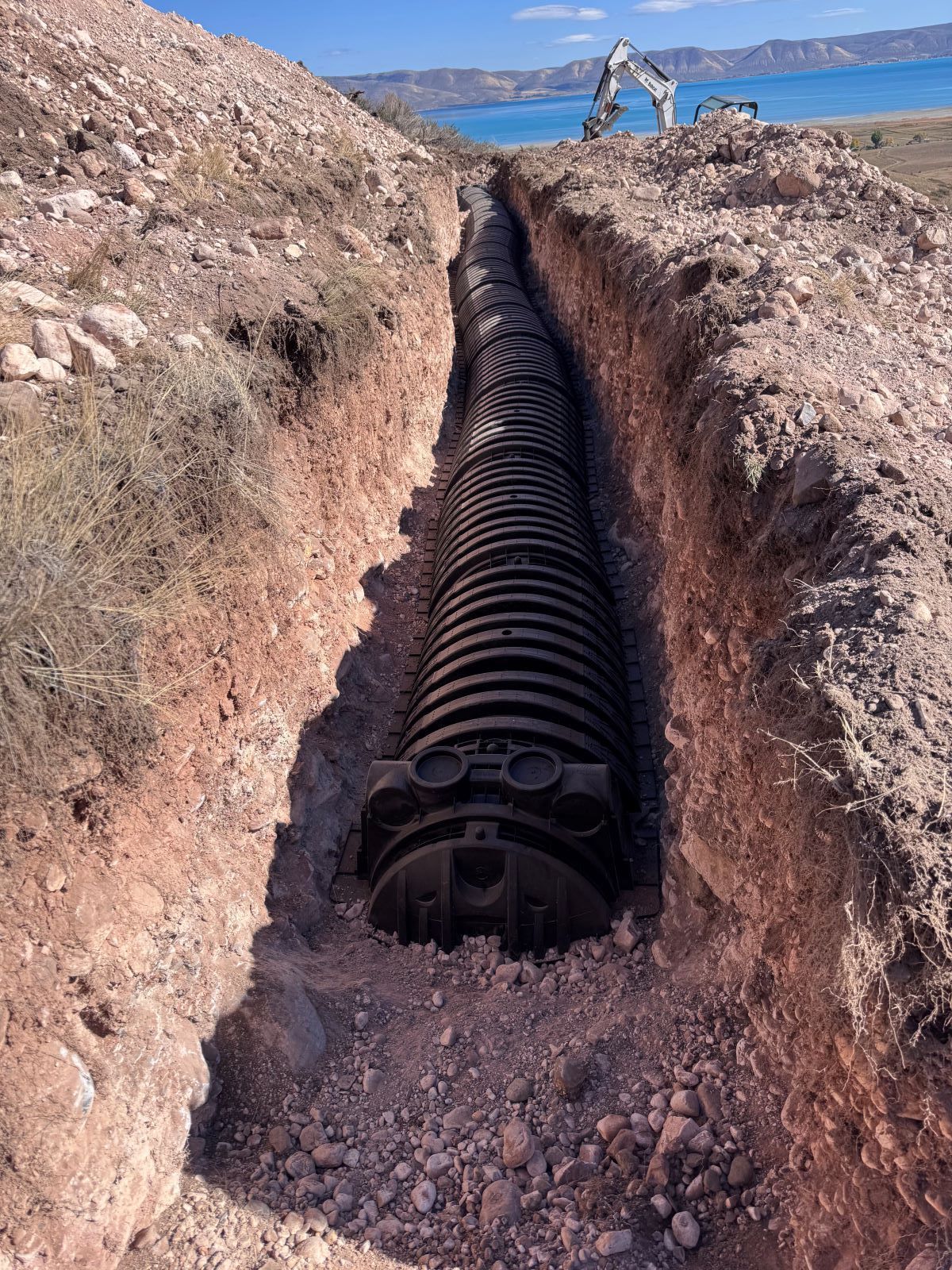 A long corrugated pipe is buried in a deep trench, likely for drainage.  An excavator is visible in the background.