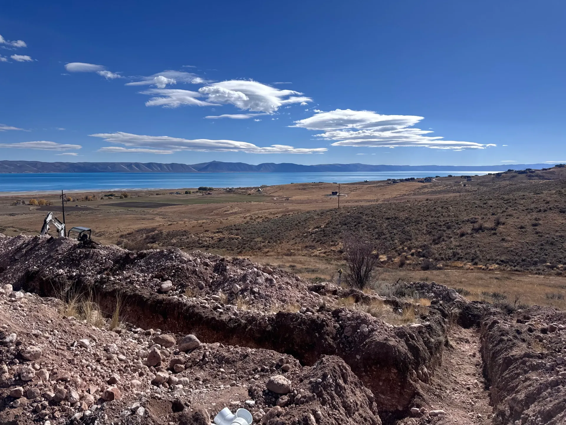Dry, brown landscape with trench in the foreground, water and blue sky in the background.