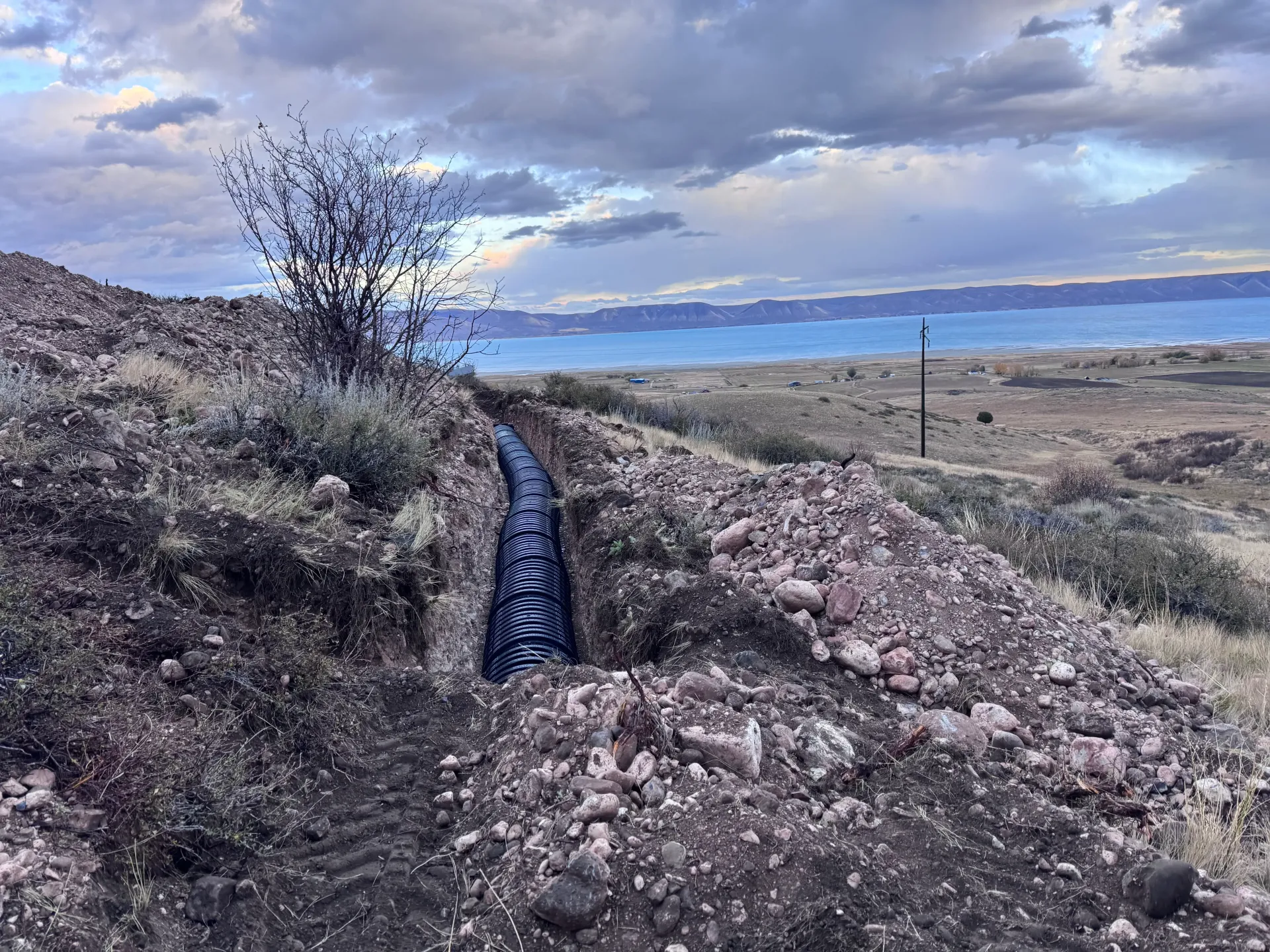 A trench with black corrugated pipe extends into a hillside, with a body of water in the background.