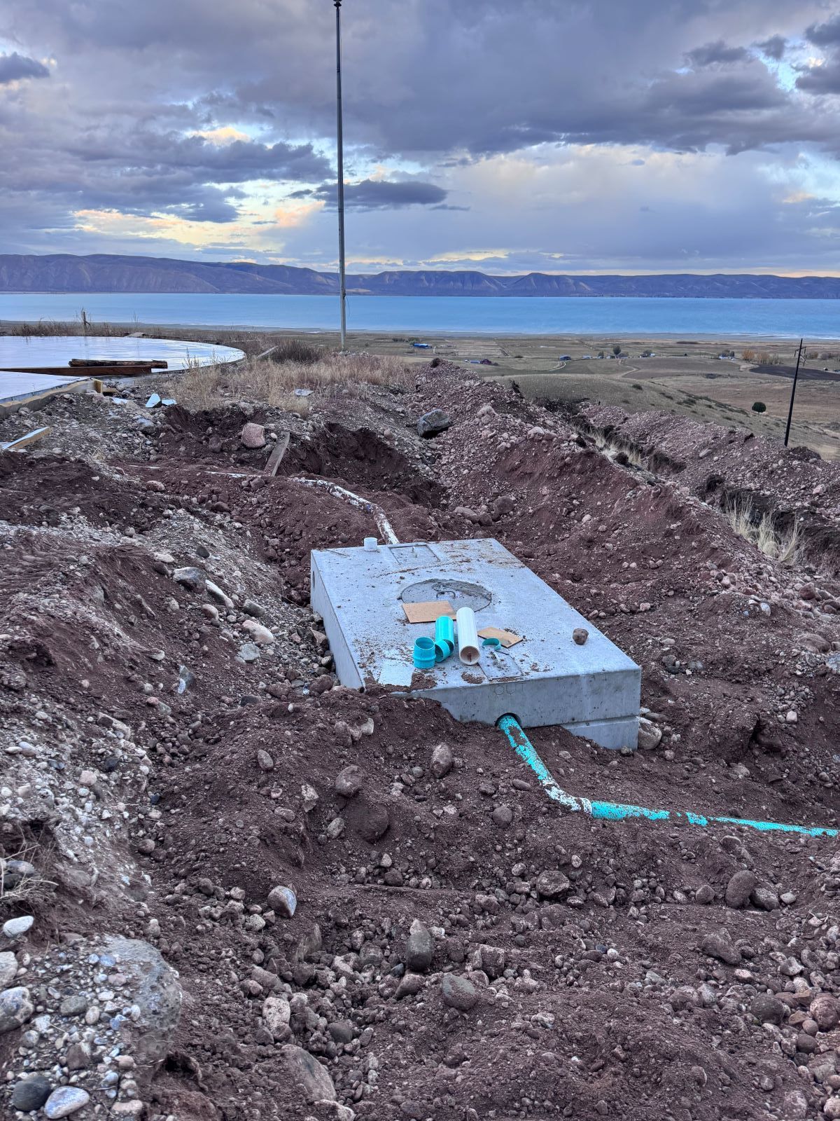 Concrete septic tank in an excavated trench, near a body of water and under a cloudy sky.