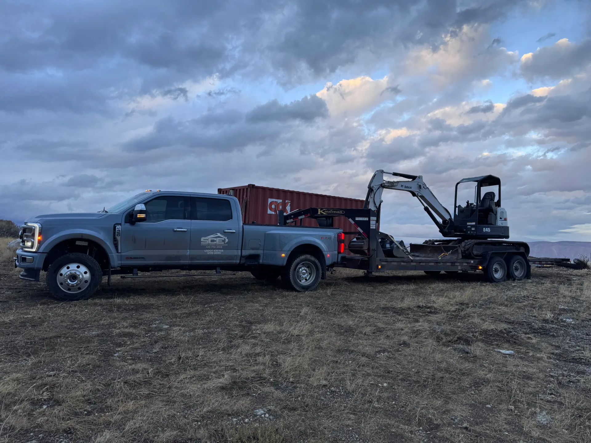 A gray Ford truck pulling a trailer with an excavator and a container on a cloudy day.