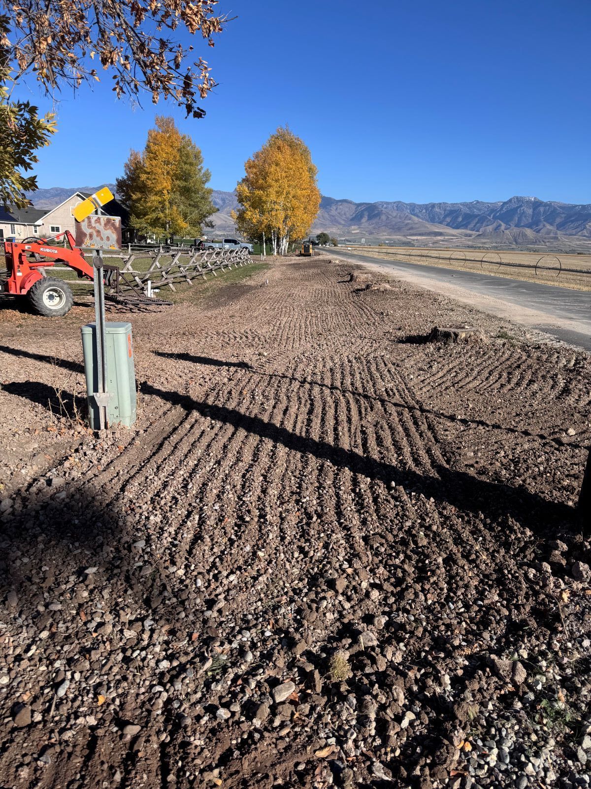 Tilled earth along a road with fall trees, mountains in the distance, and tractor on the left.