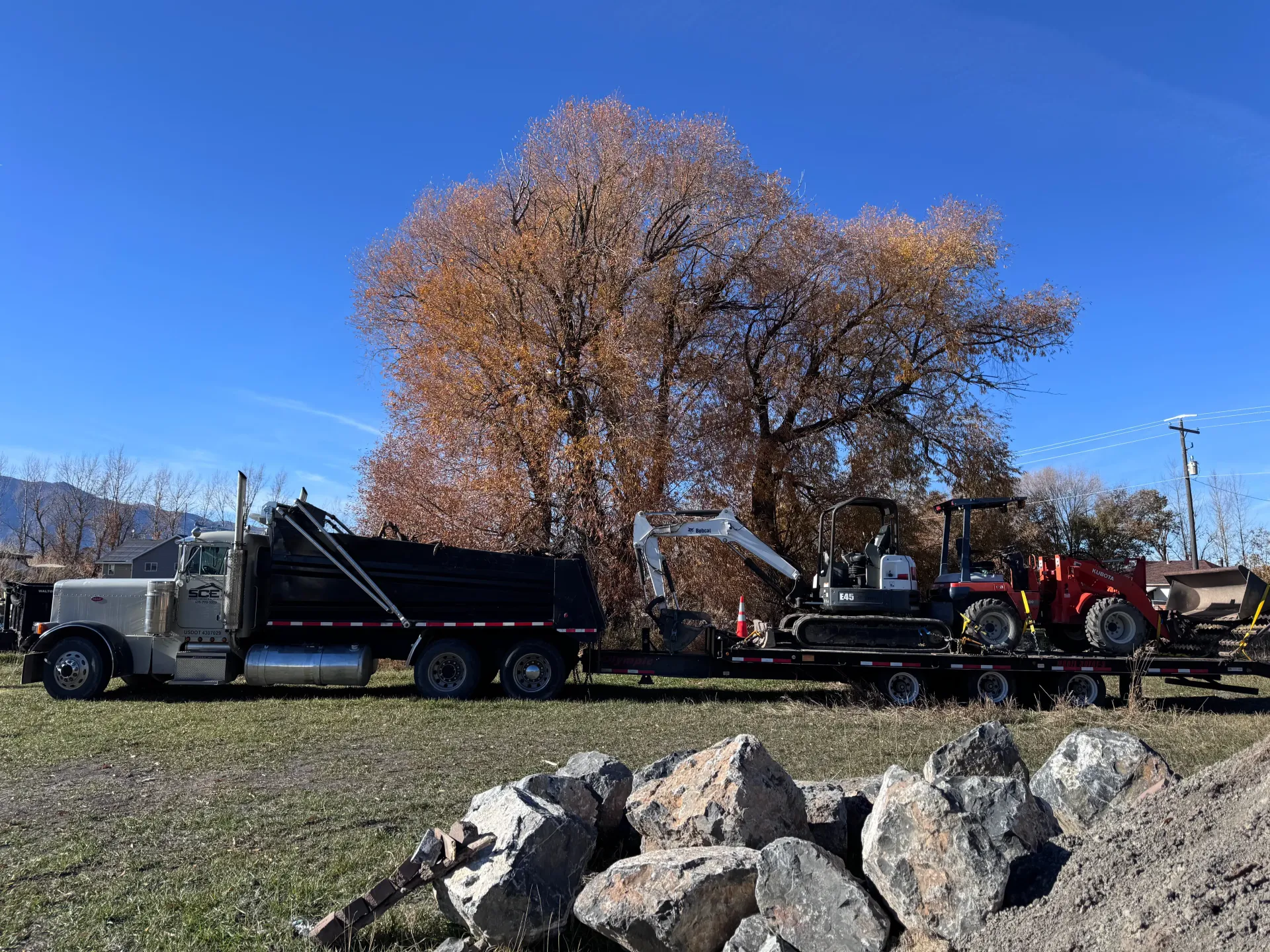 Truck with trailer carrying machinery, in front of a tree with brown leaves, under a blue sky.