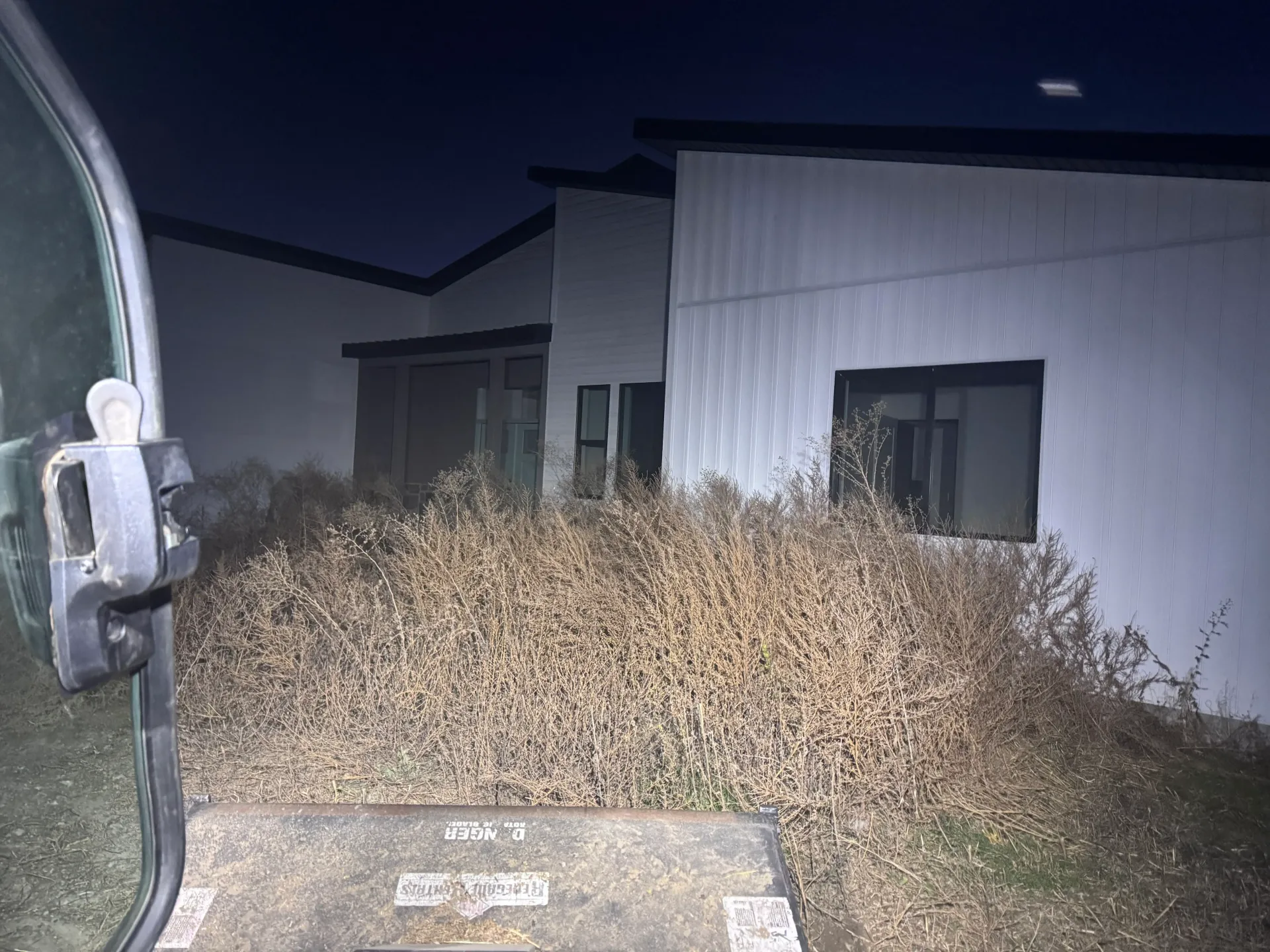Exterior of a white building at night, overgrown with dry brown plants. Part of a vehicle is in the foreground.