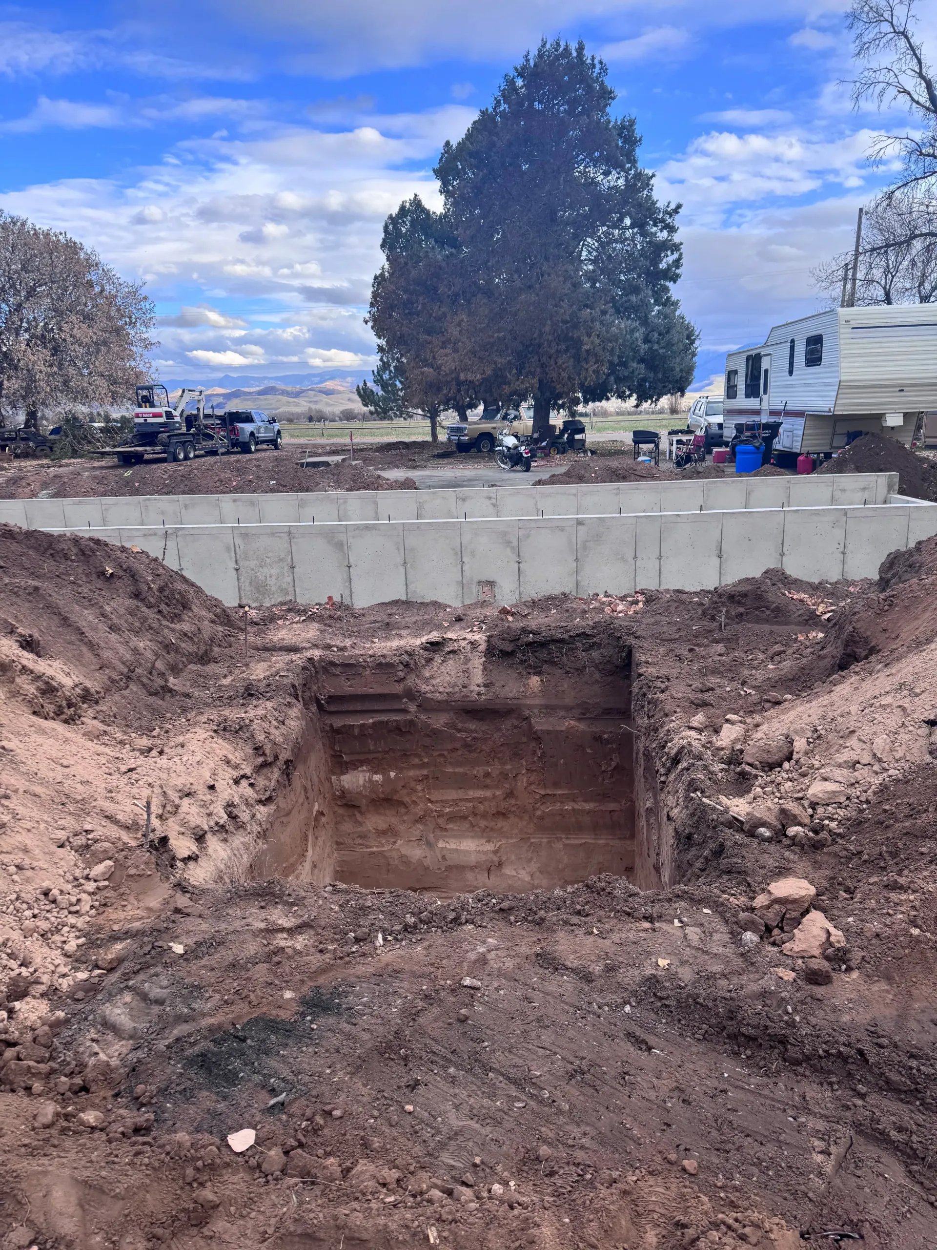Excavation pit in front of a concrete foundation under construction; tree and cloudy sky in background.