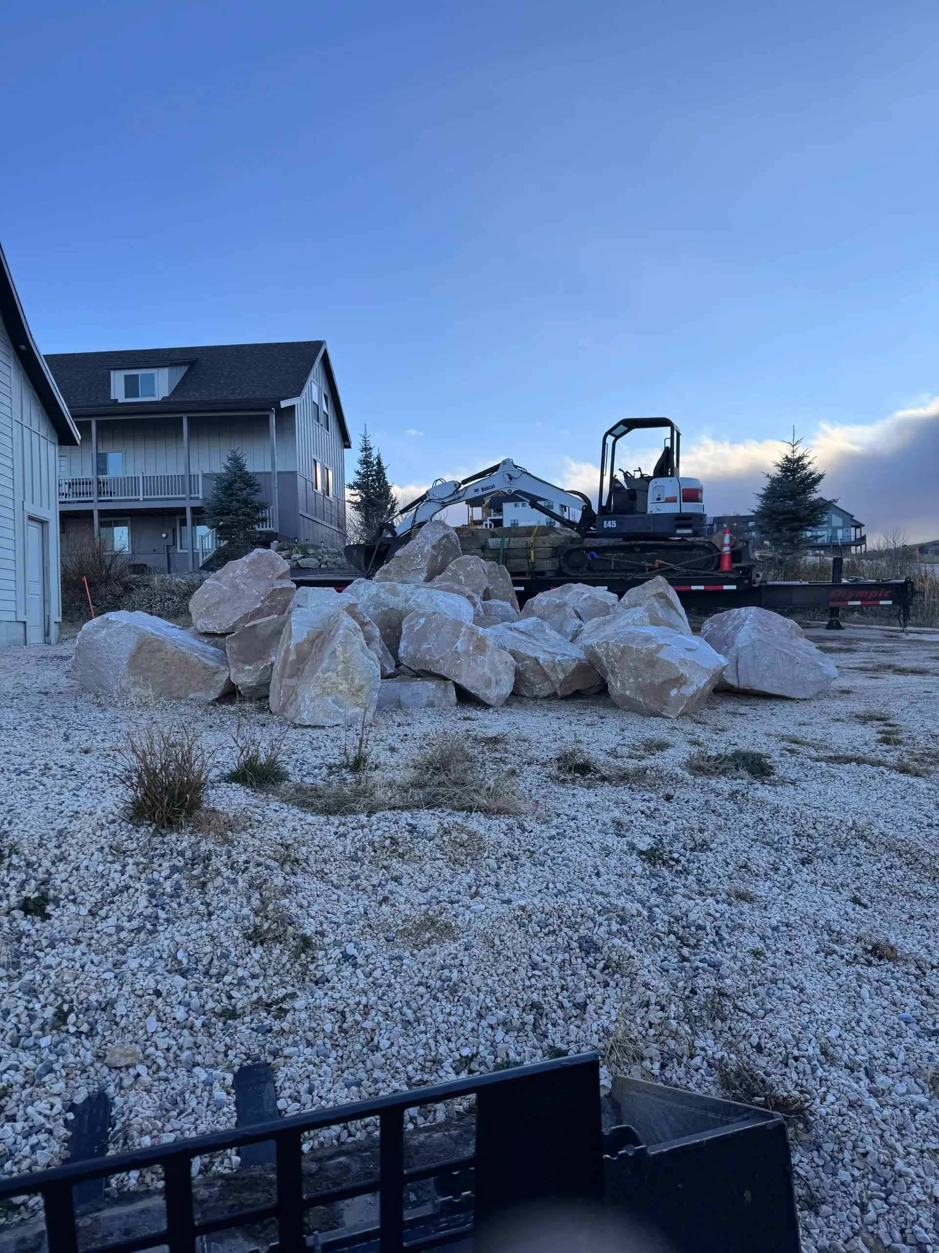 Pile of large rocks on a gravel yard with a small excavator and houses in the background under a blue sky.