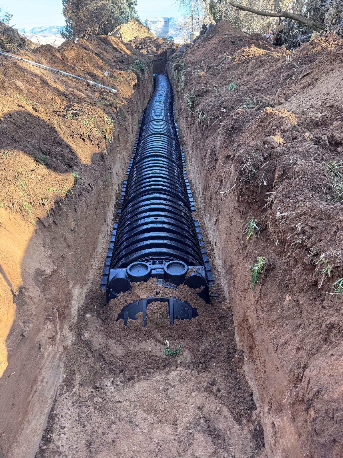 A long, narrow trench containing black drainage cells, surrounded by brown soil, outdoors.
