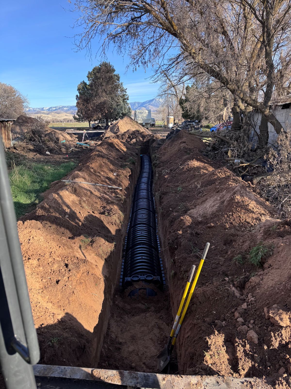 Trench with black drainage system installed, shovel nearby. Brown soil, blue sky, and trees.