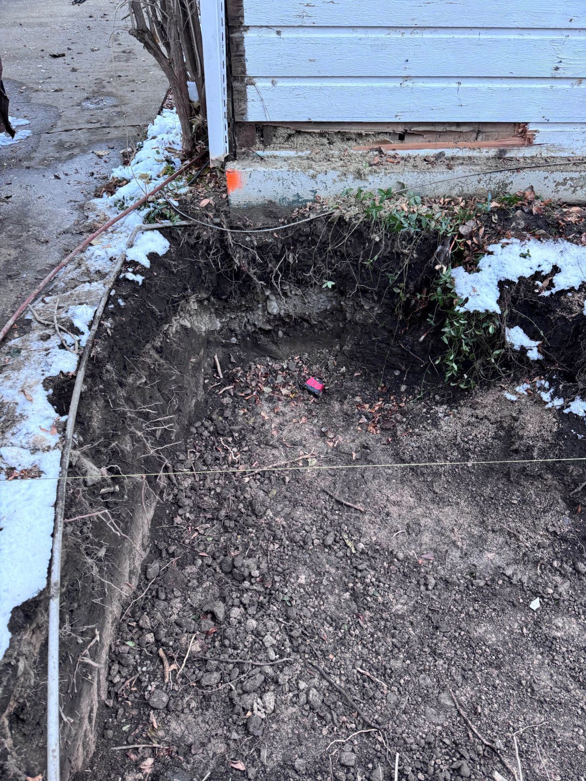 Excavated dirt bed next to a sidewalk and building with snow patches.