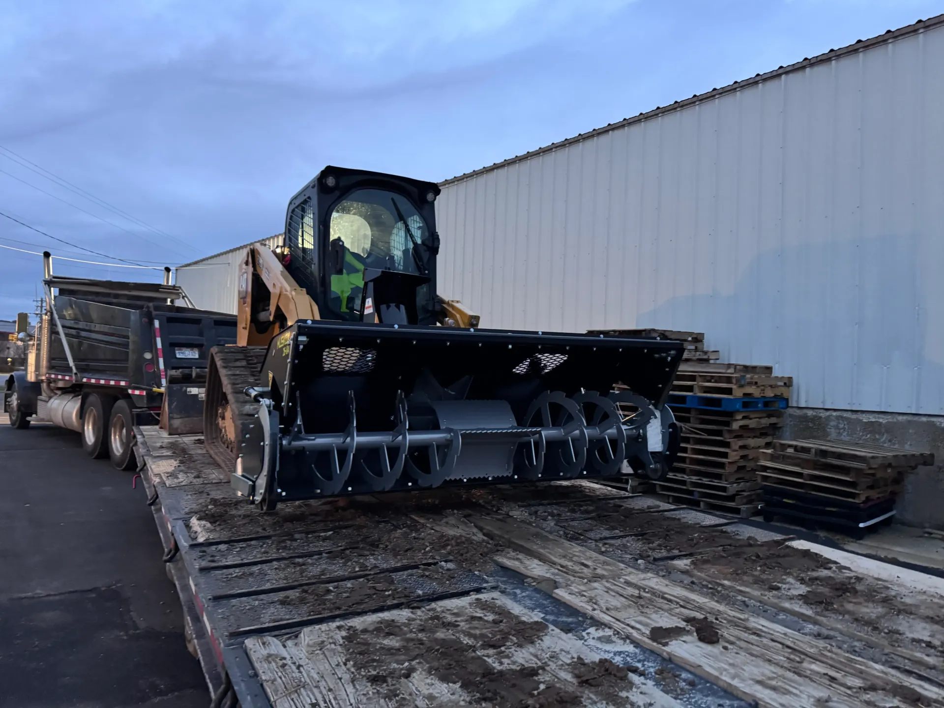 Skid steer with snowblower attachment on a flatbed trailer in front of a building.