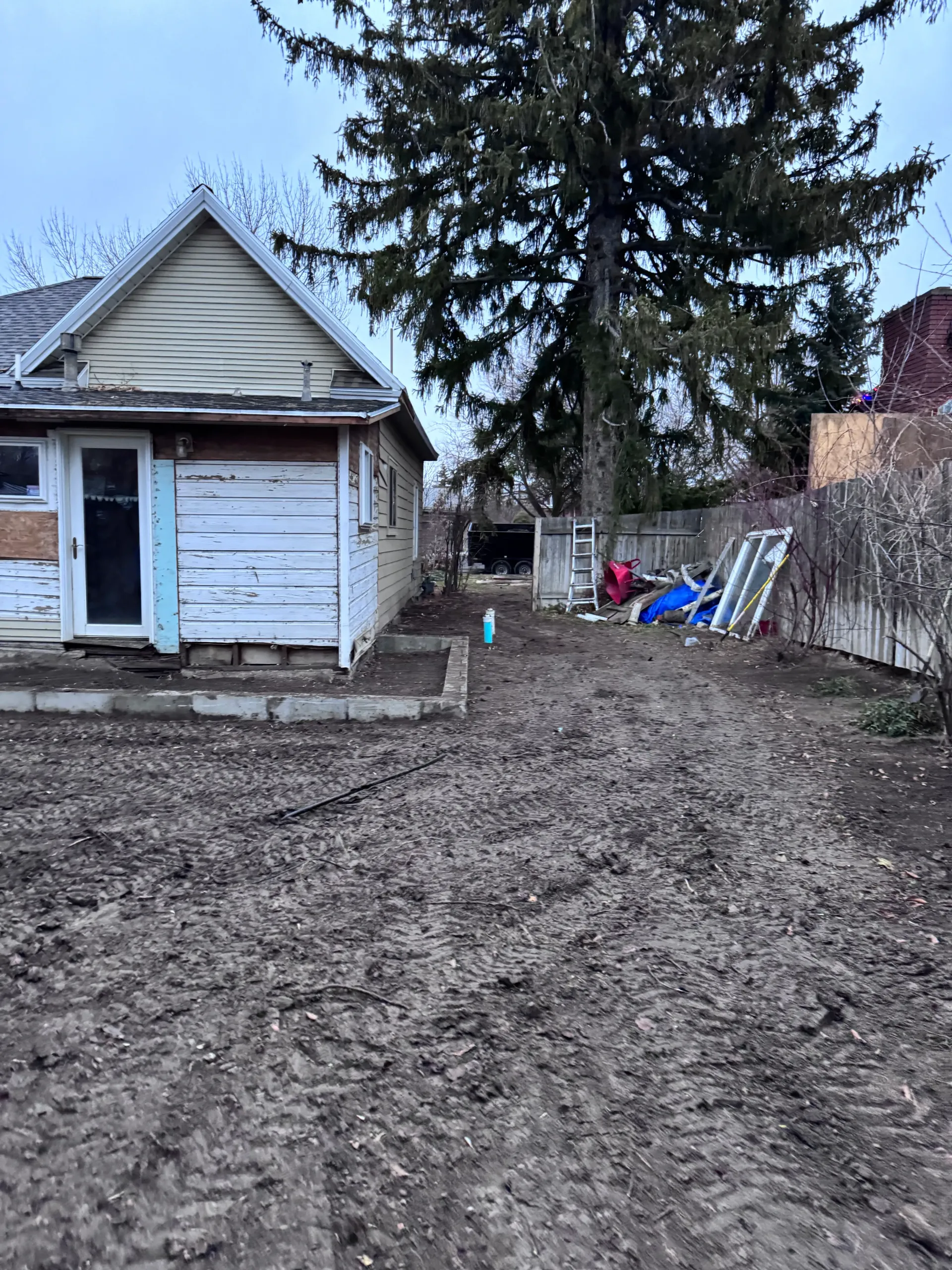 Dirt driveway next to weathered garage with peeling paint. Tall tree, wooden fence, and scattered debris are present.
