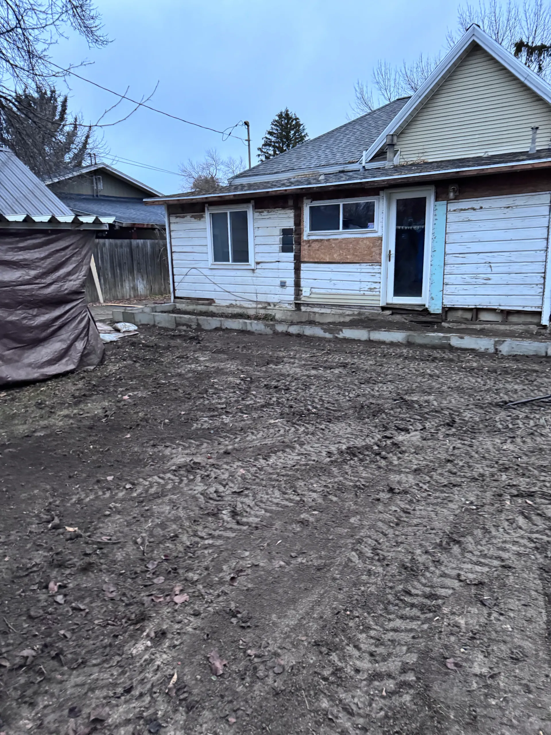Exterior of a weathered, single-story house with a dirt yard, under an overcast sky.