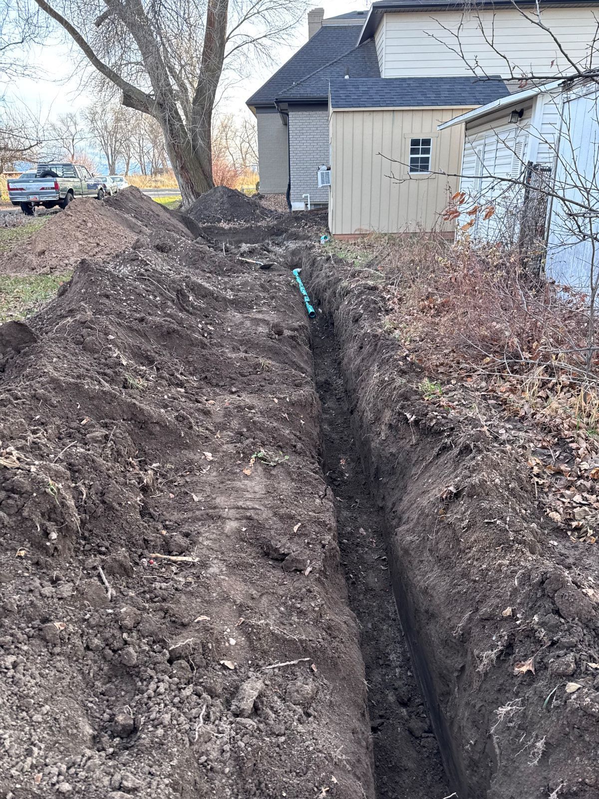 A long trench dug in a yard with a blue pipe visible; a house and shed are in the background.