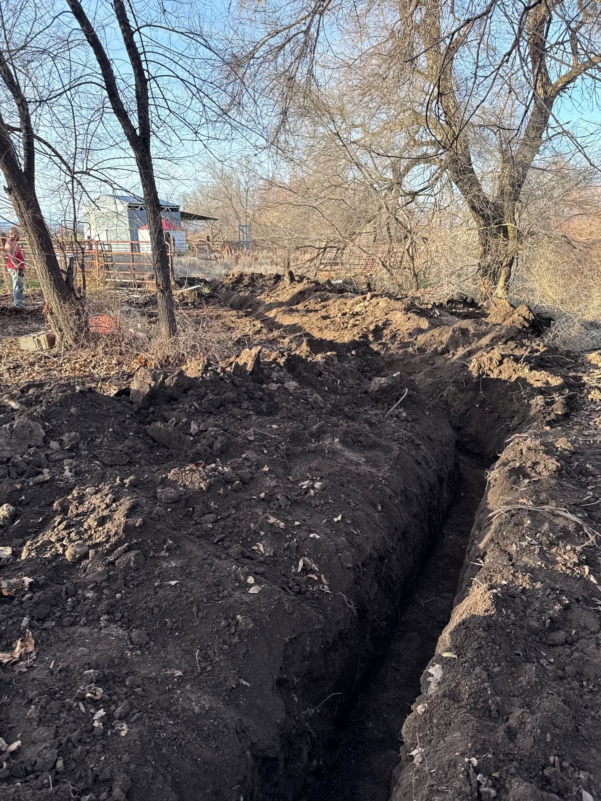 A deep trench dug in dark soil near trees and a building in the background.