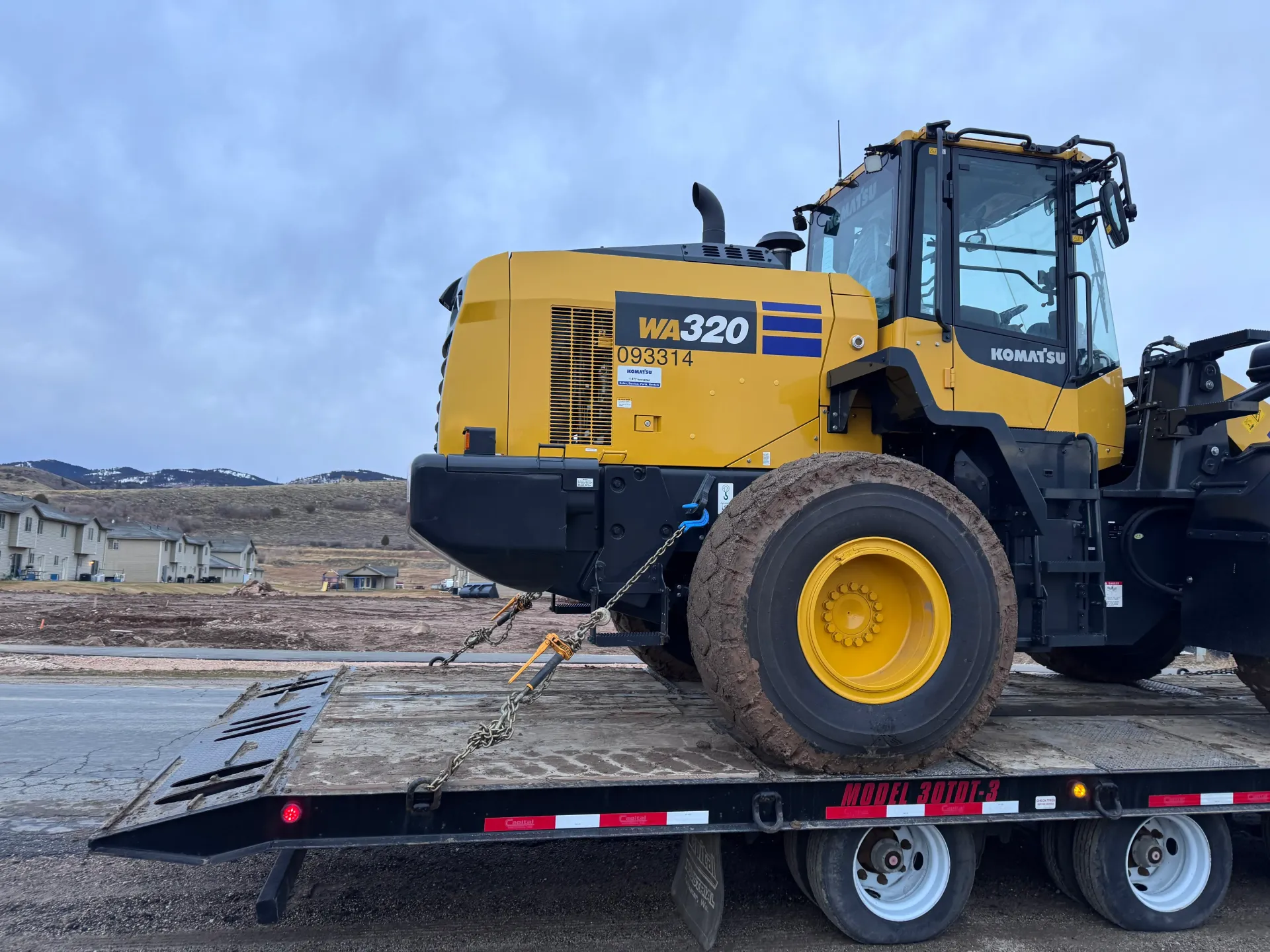 Yellow Komatsu WA320 wheel loader on a flatbed trailer, overcast sky in background.