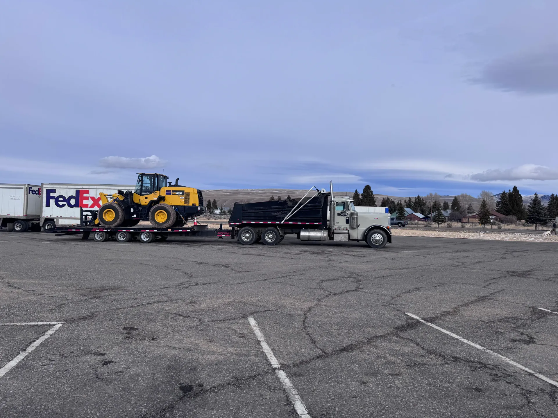 A semi-truck hauling a Caterpillar front loader and a dump trailer in a parking lot under a cloudy sky.