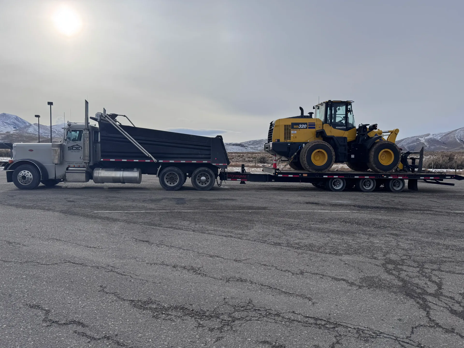 Grey semi-truck towing a black dump trailer and a yellow front-end loader on a flatbed trailer; parked on pavement.