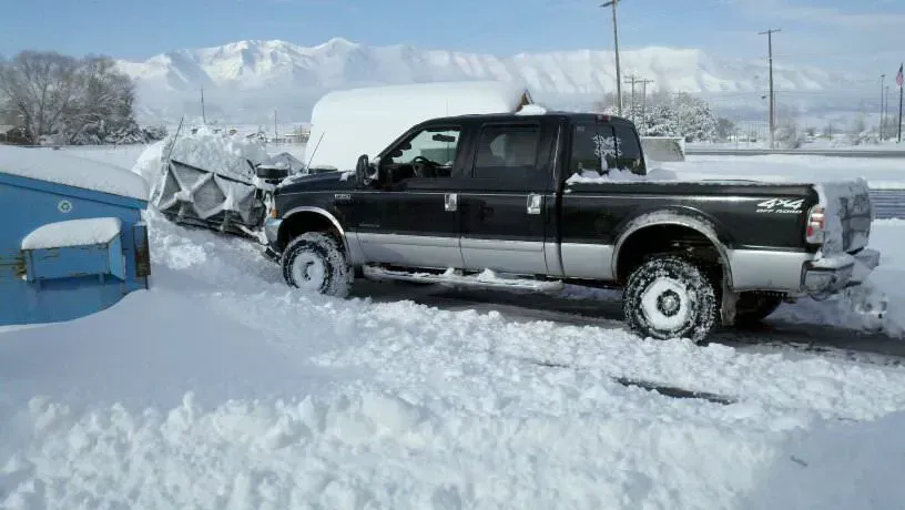 Snowplow truck plowing snow in front of a blue storage container; snow-covered mountains in the background.