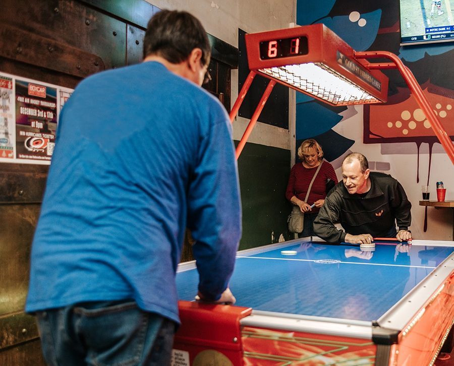 People playing air hockey in a bar. Man in blue shirt facing the game, two others watching, score 6-1.