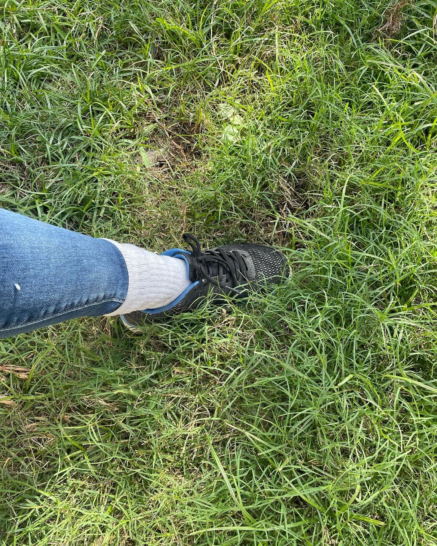 A person's foot in a dark shoe, blue jeans and a white sock on green grass.