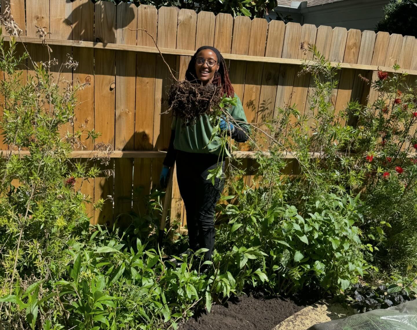 Woman gardening in front of a wooden fence, holding branches, smiling. Green plants and sunny day.