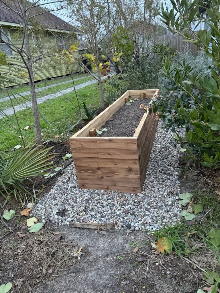 Wooden raised garden bed on gravel, in a yard with plants and a path.