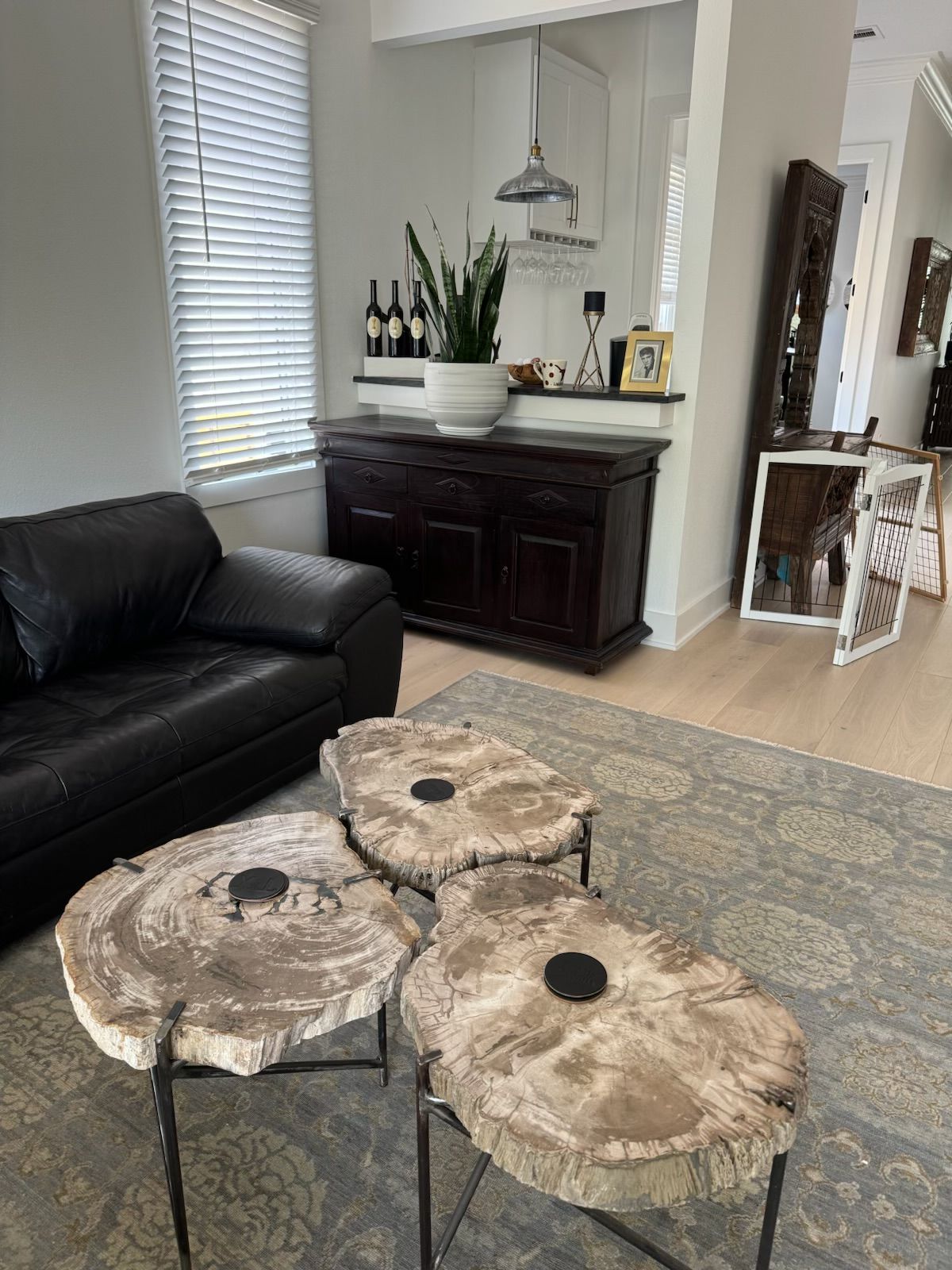 Three wood slab coffee tables on rug, black leather sofa, bar with plants and wine.