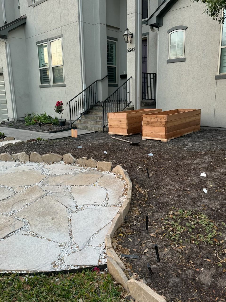 Two wooden planters sit on a brown soil yard, next to a stone pathway and a white house.