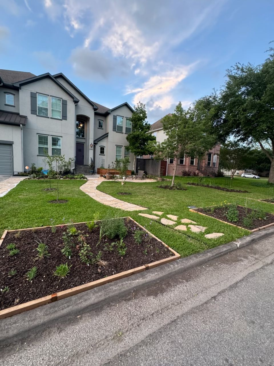 Two-story light gray house with stone walkway and landscaped yard on a suburban street.