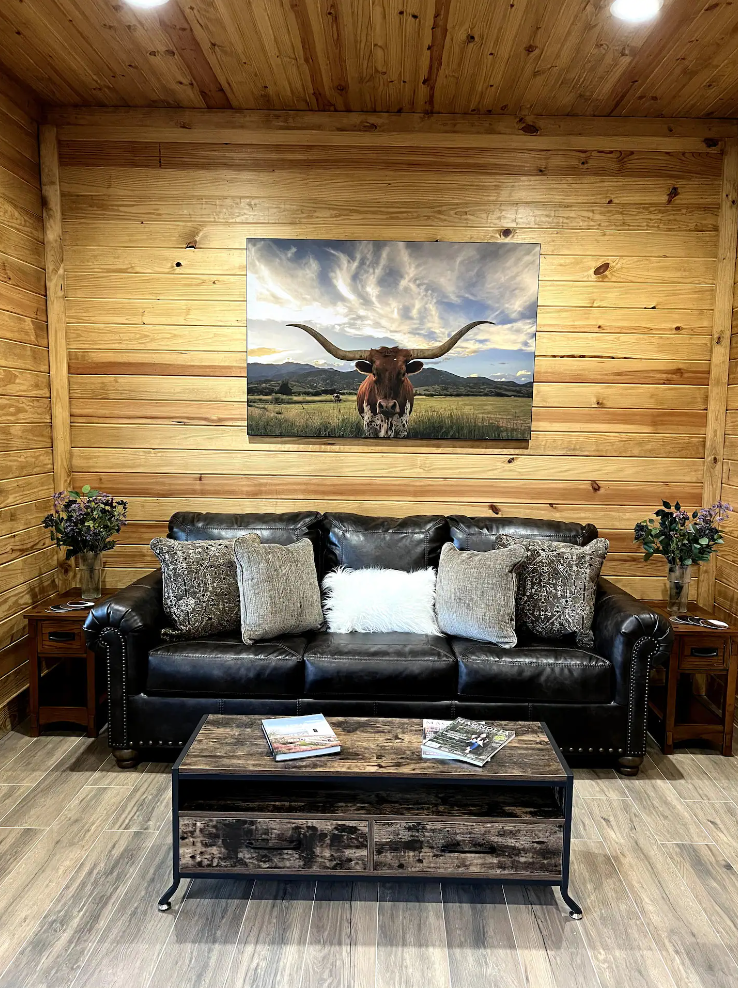 A living room with a couch , coffee table , and a painting of a longhorn on the wall.